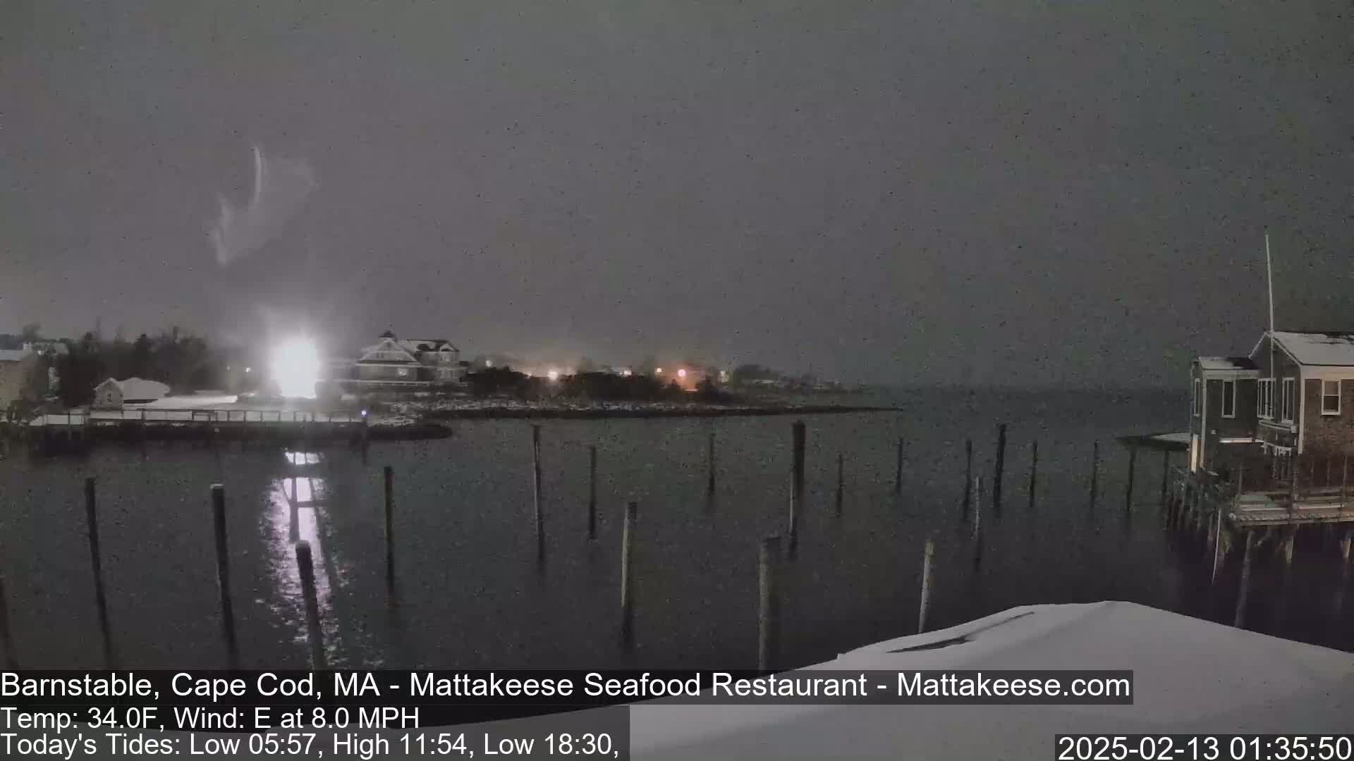 A nighttime, snowy scene shows a calm body of water with posts extending from the shore, buildings in the background, and a faint light in the distance under a dark, overcast sky.