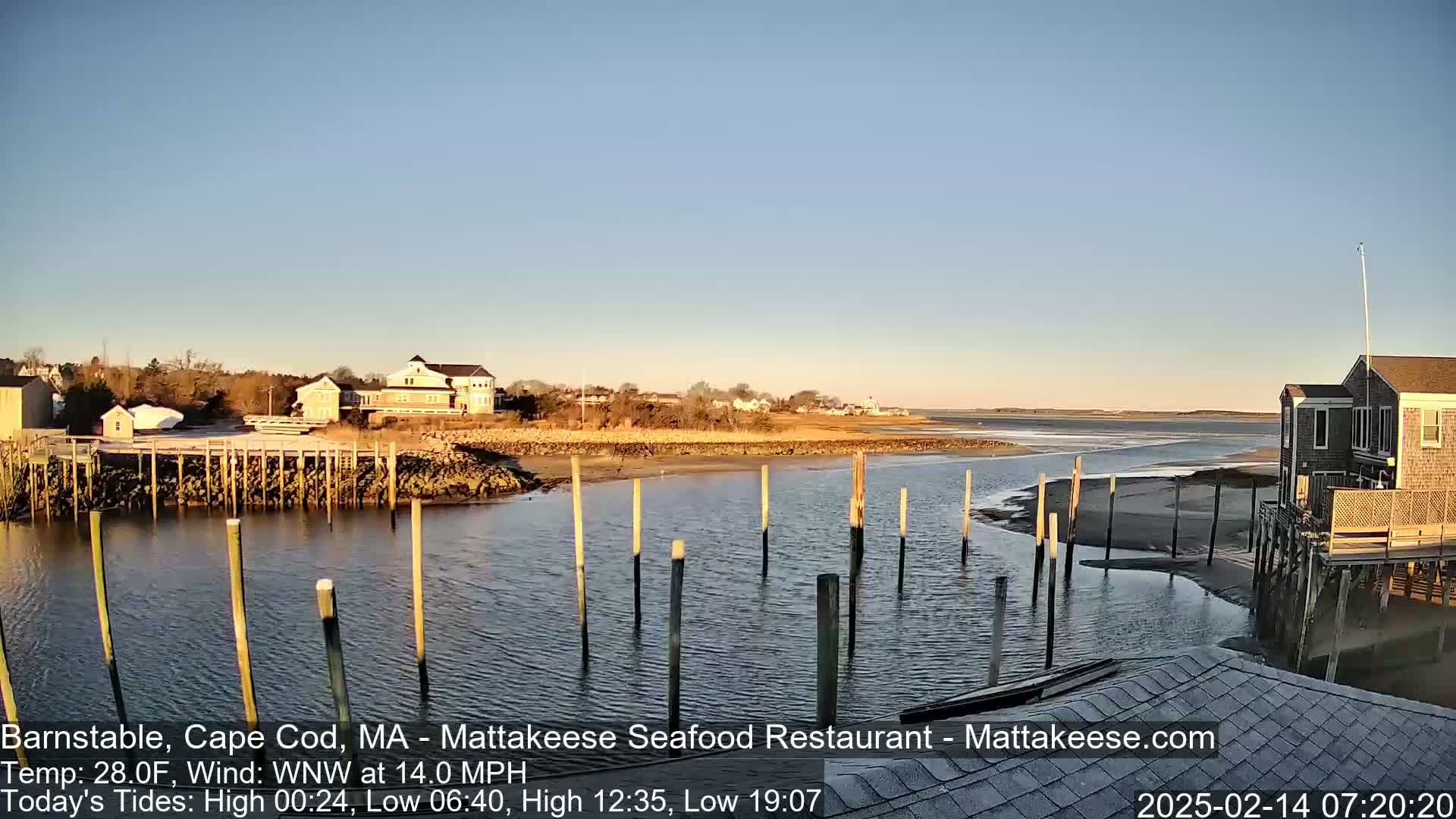 A calm, clear day shows a waterfront scene with several buildings, a small dock, and calm water under a mostly clear sky.