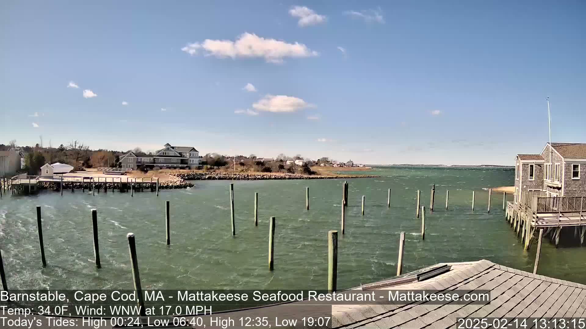 A choppy body of water with wooden pilings extends from a shoreline with buildings, under a partly cloudy sky.