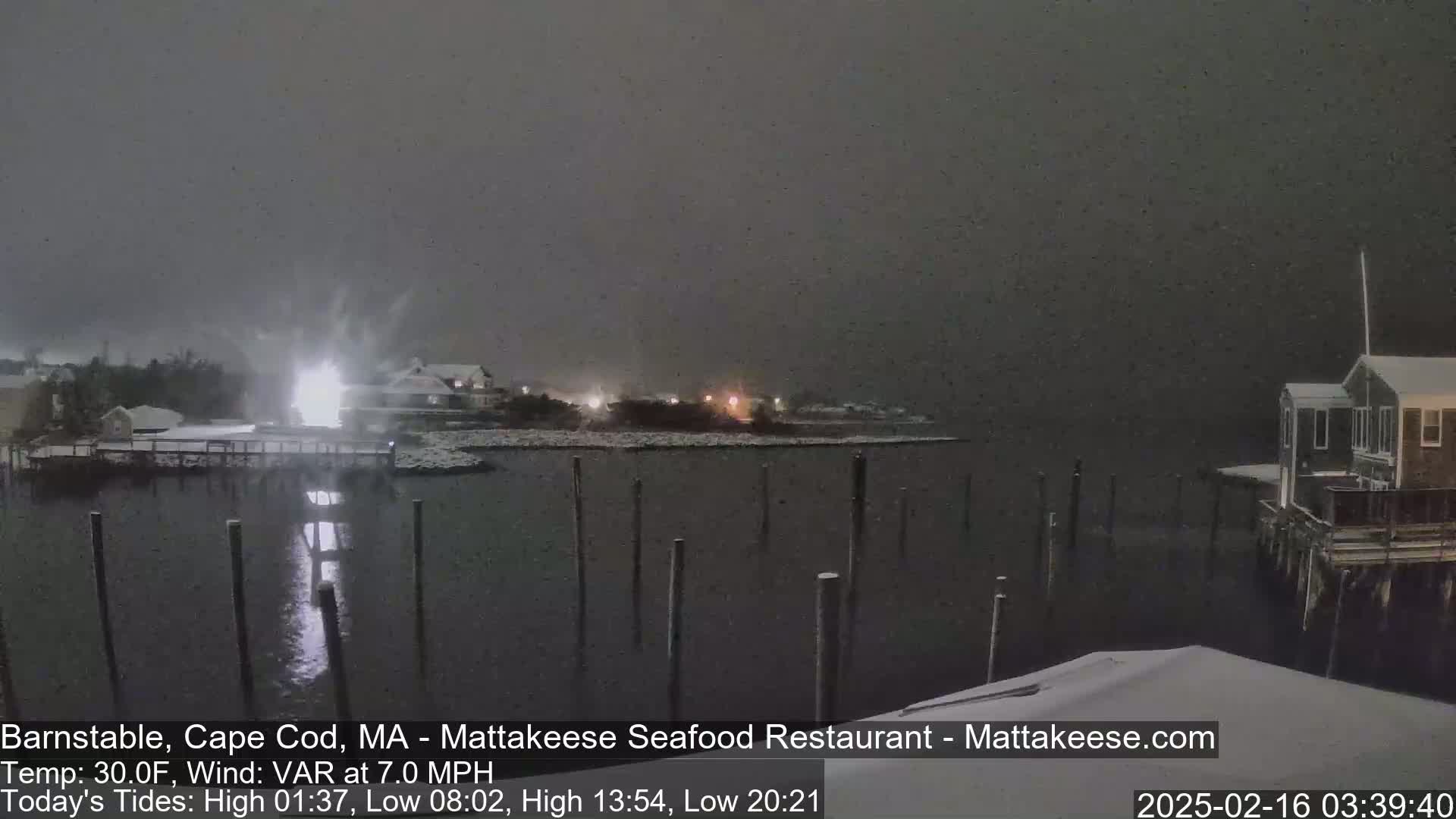 Snow falls on a dark, waterfront scene with snow-covered buildings and a calm body of water containing many posts.