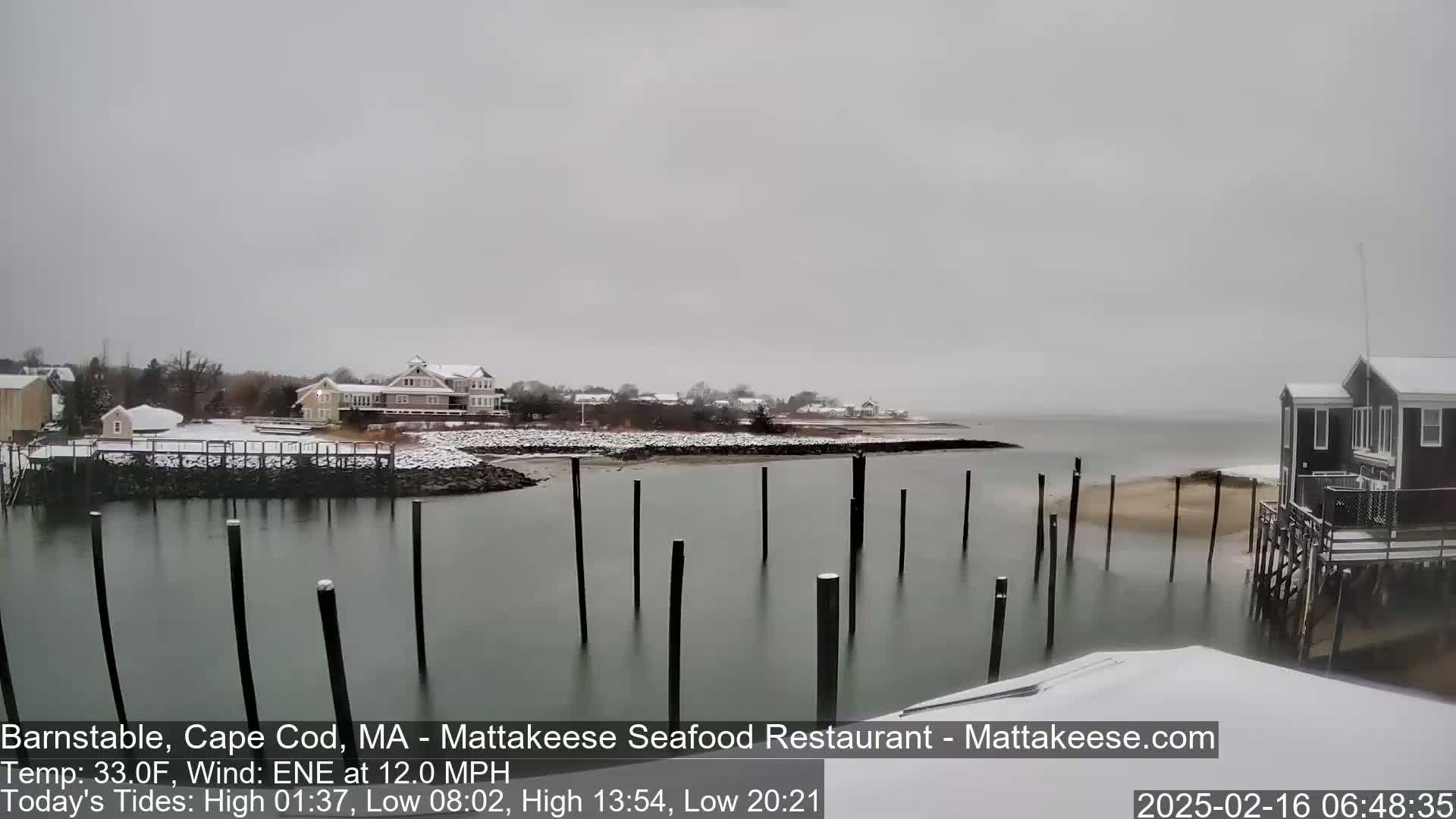 A snow-covered waterfront scene shows a calm body of water with several pilings, houses on a snow-dusted shore, and a light snowfall.