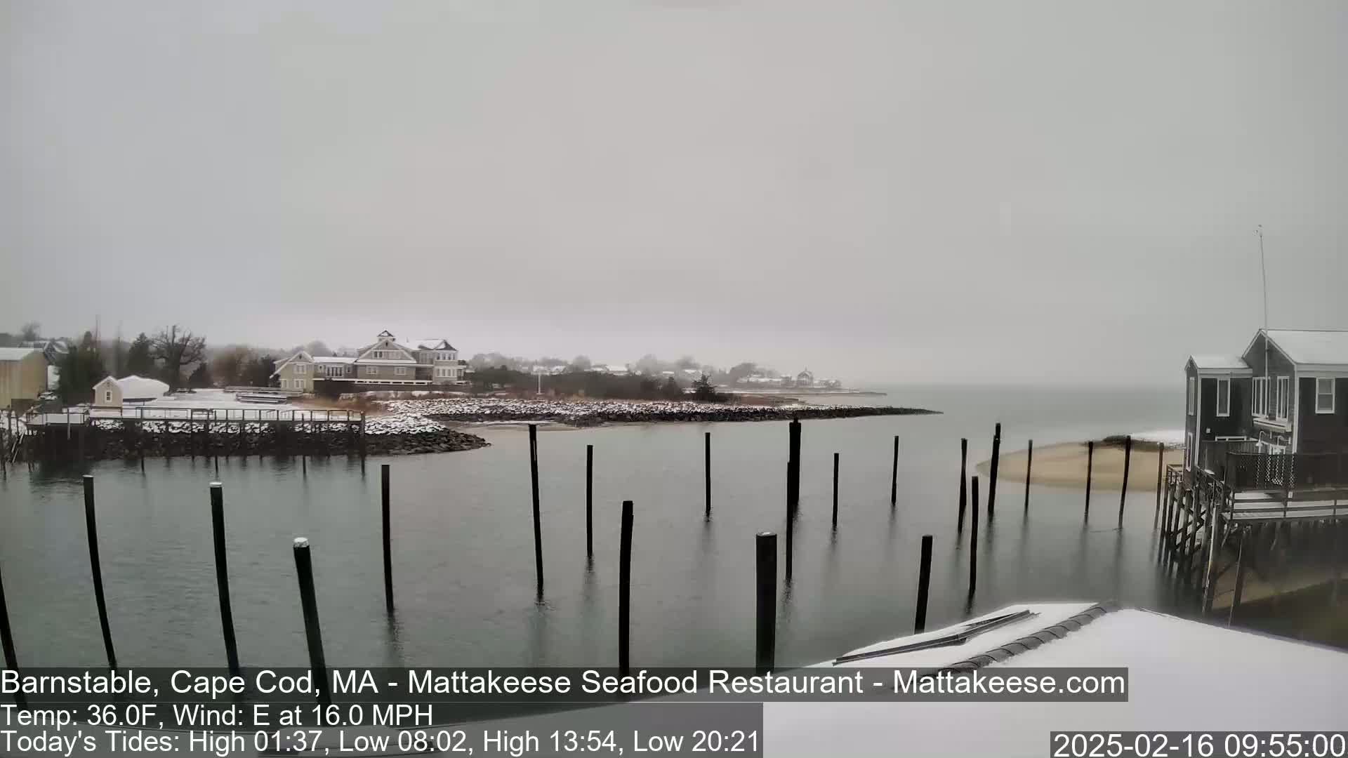 A snow-covered waterfront scene shows a calm body of water with wooden pilings, a few buildings, and a light dusting of snow on the ground.