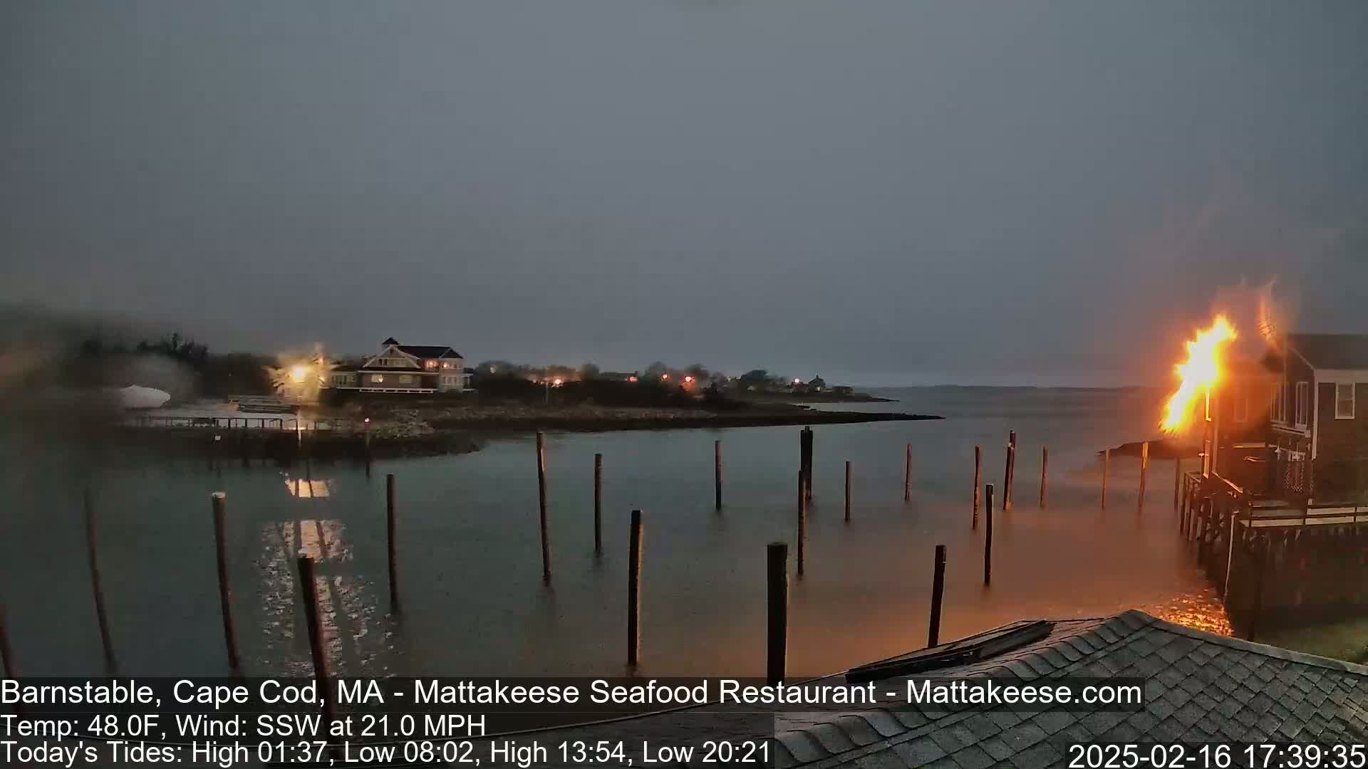 A waterfront building is on fire under a dark, overcast sky with a strong wind.