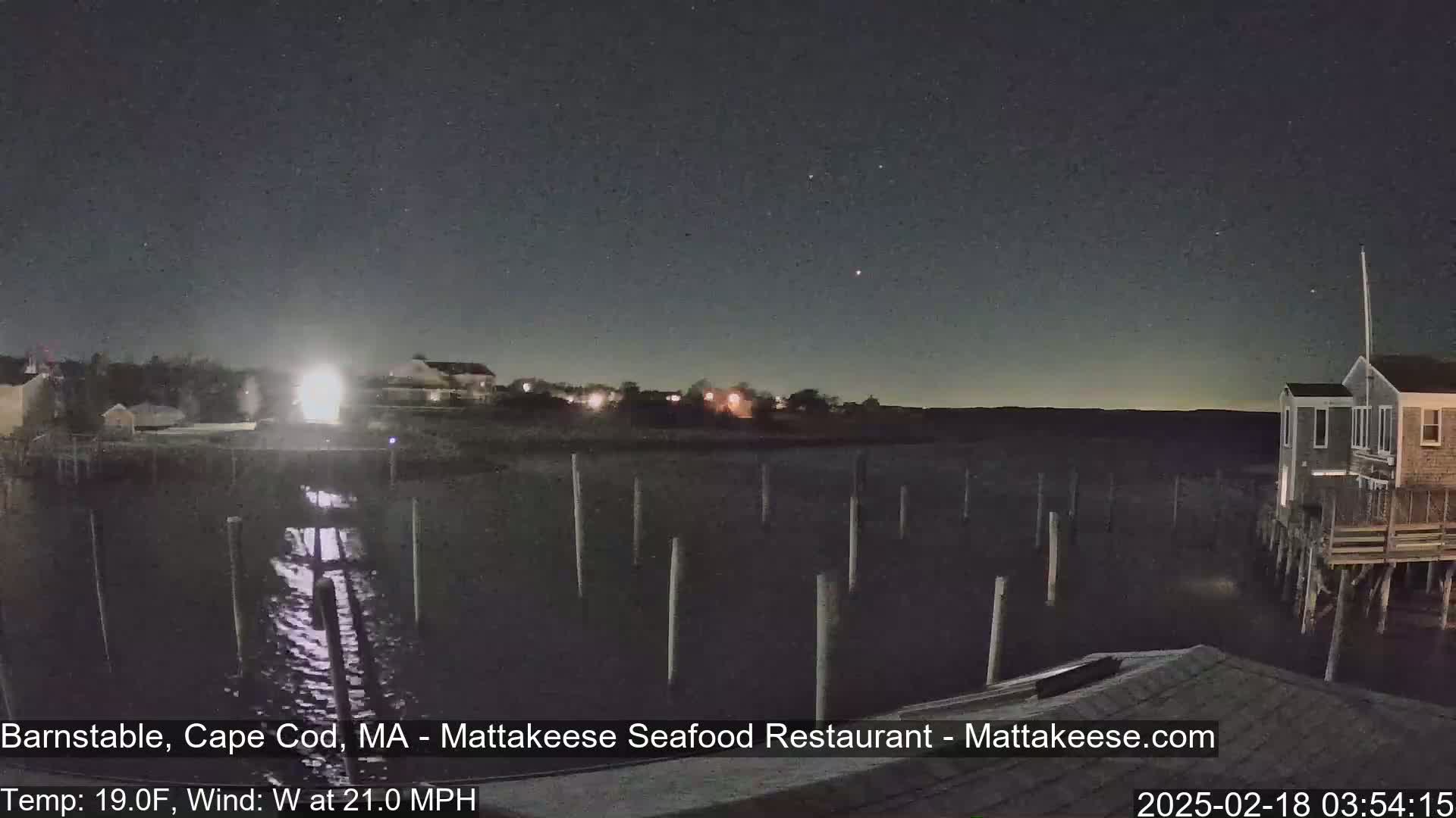 A nighttime view of a calm body of water with pilings, a few buildings, and a starry sky, under a 19°F temperature with a 21 mph westerly wind.