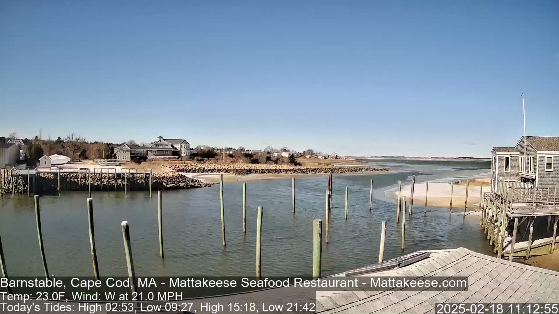 A calm, sunny day reveals a waterfront scene with a small harbor, houses, and a restaurant on stilts, under a mostly clear blue sky.
