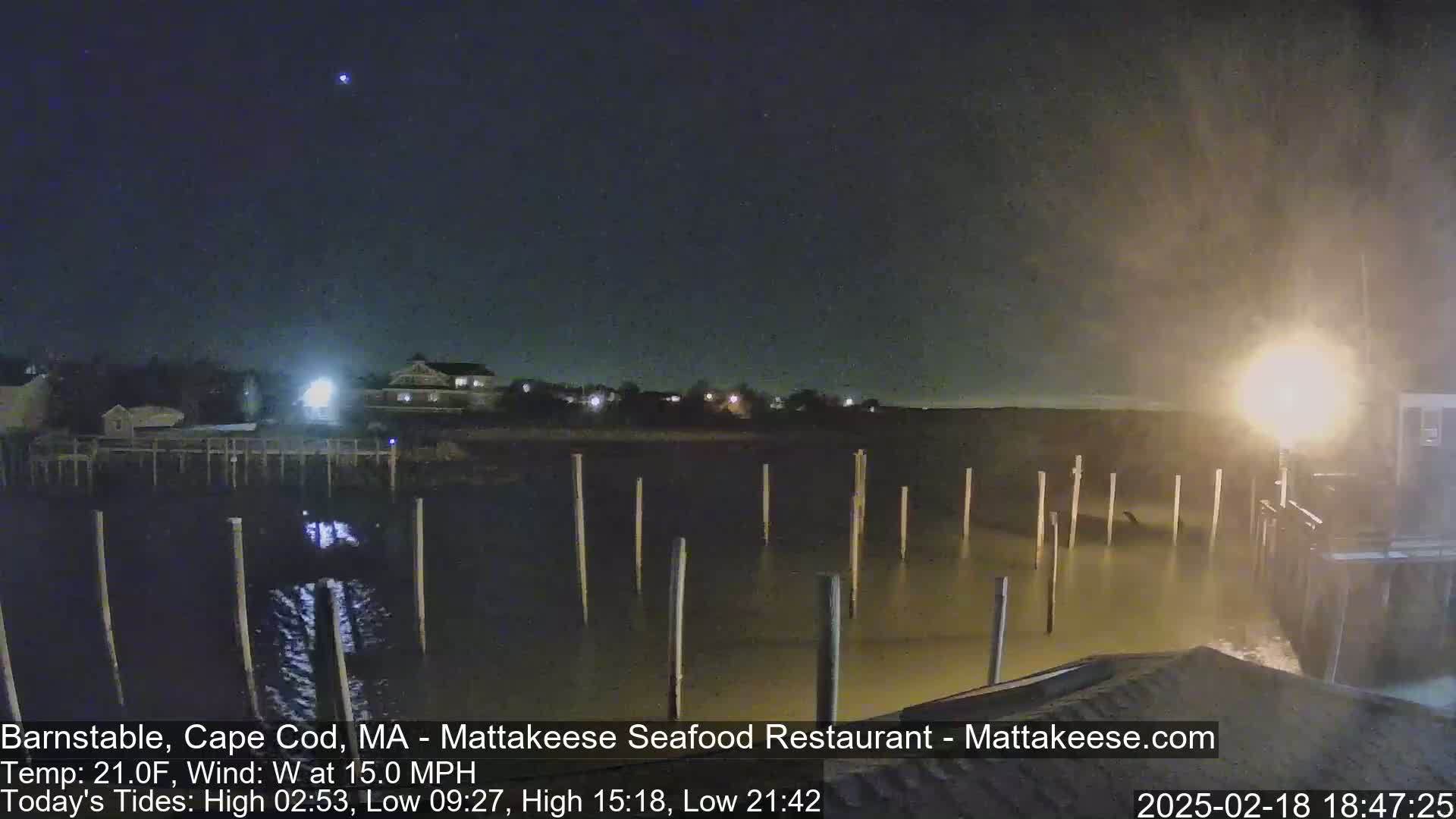 A nighttime view of a calm body of water with wooden pilings extending into it, under a dark sky with a few visible stars and a strong west wind.