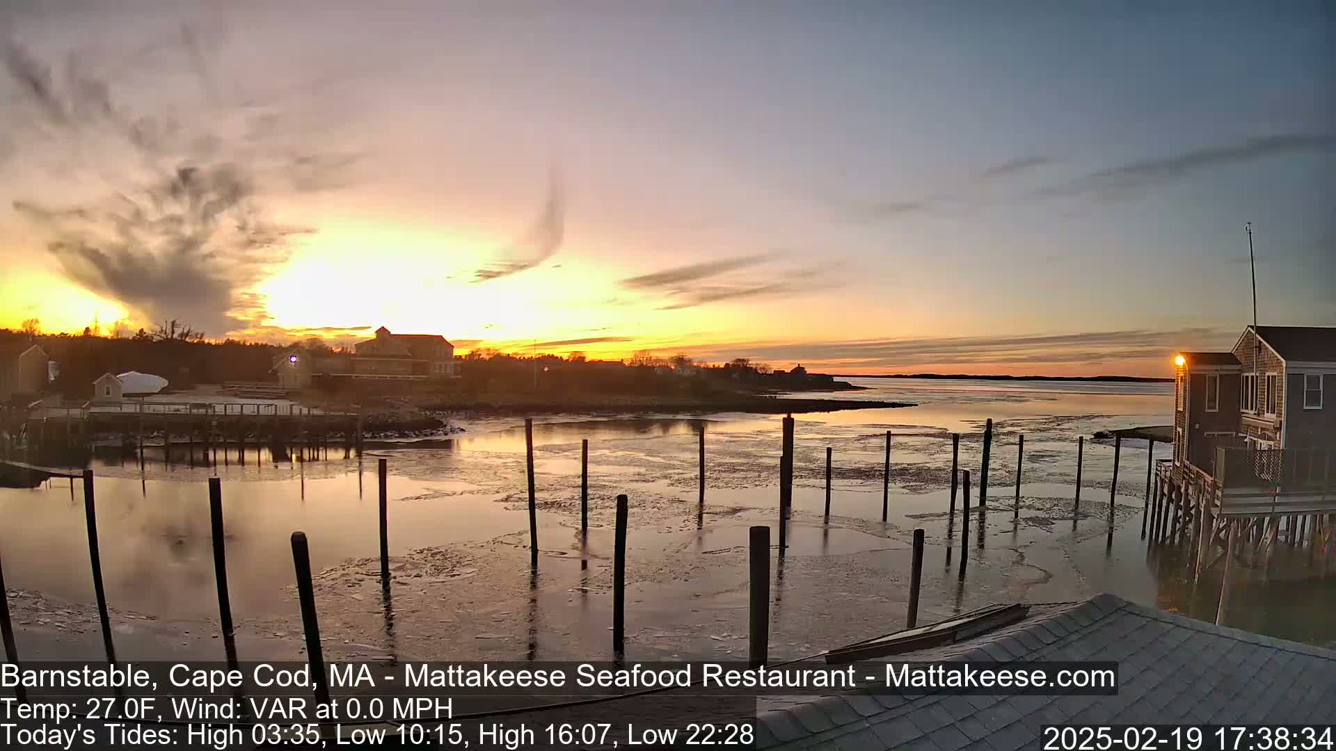 A calm waterfront scene at sunset with partially frozen water, showing a few buildings and pilings.