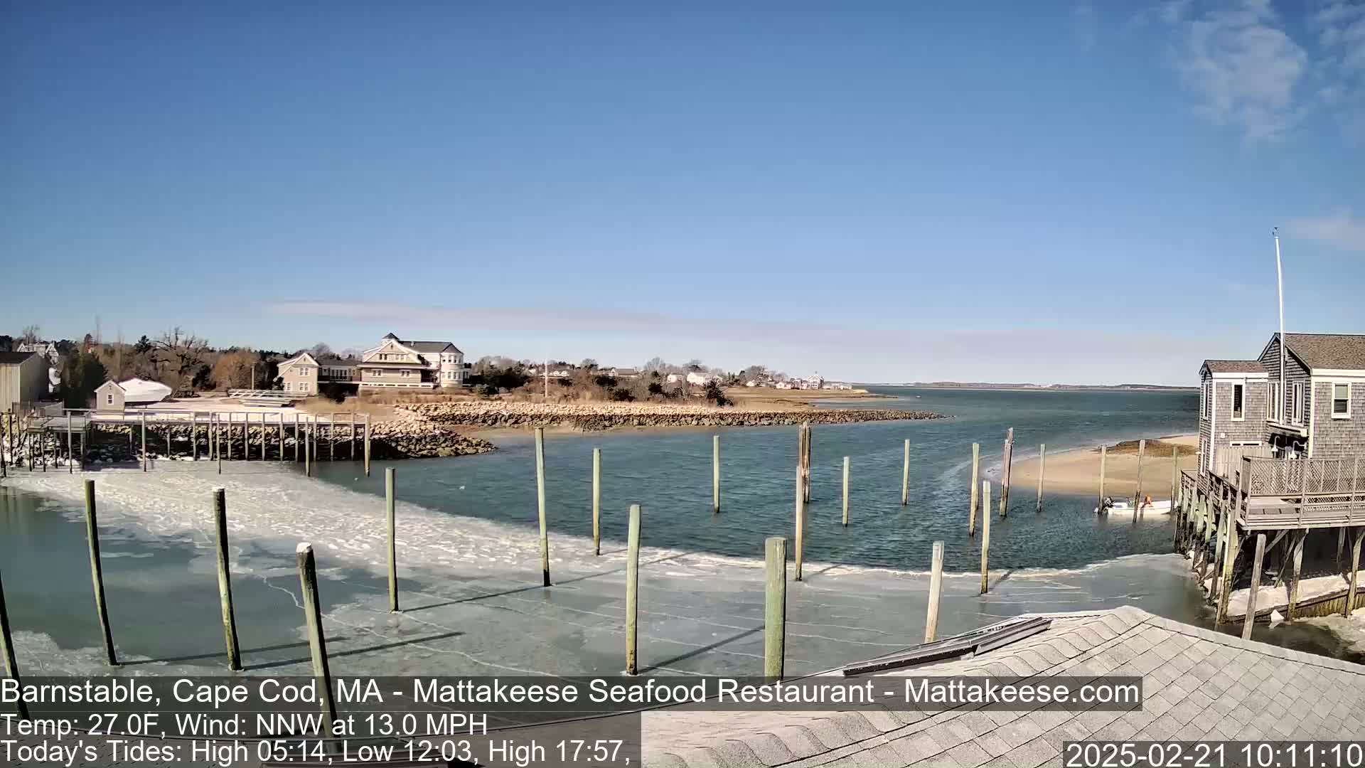A partially frozen harbor with wooden pilings, buildings on a shoreline, and a clear blue sky.