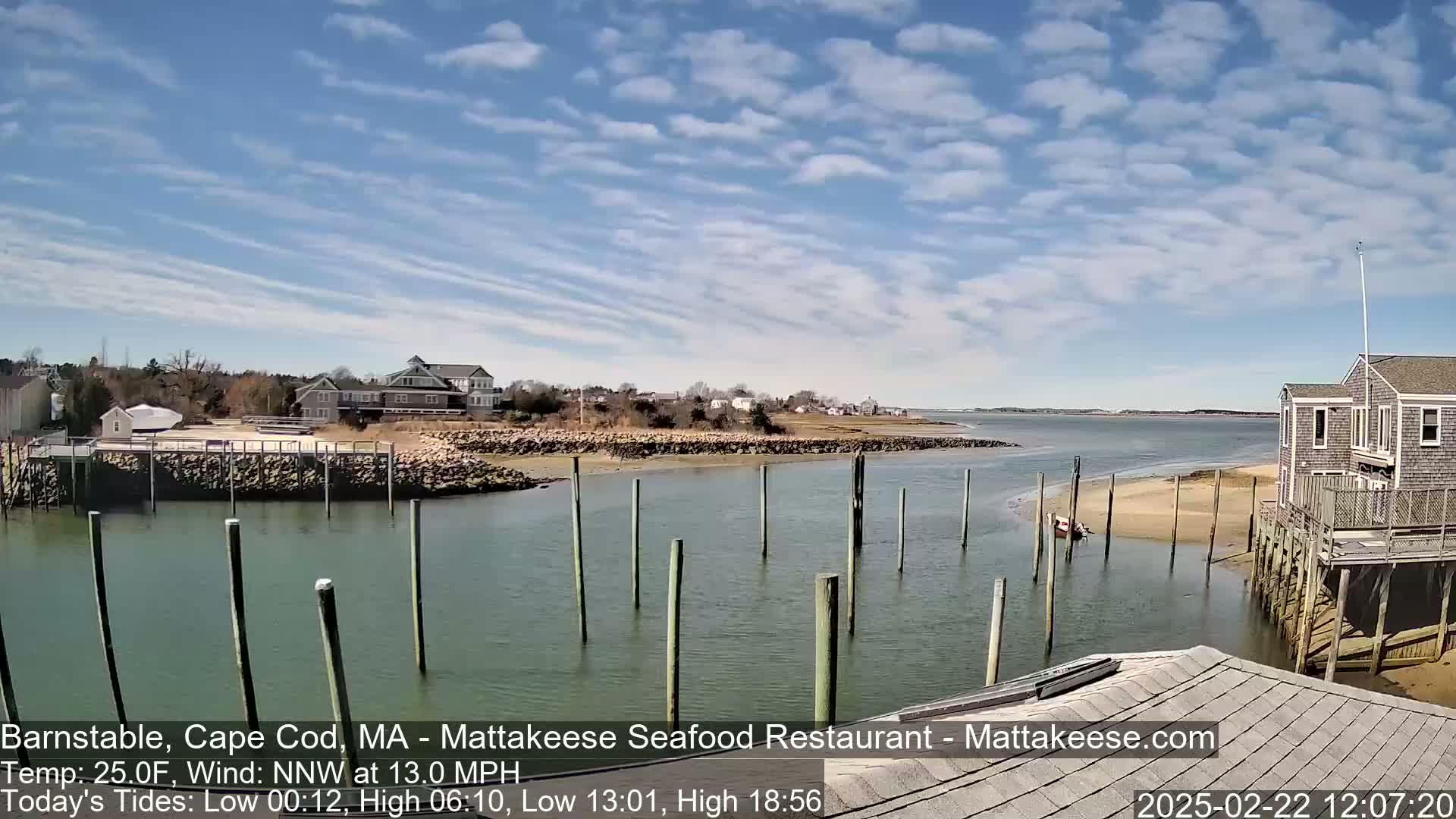 A partly sunny day shows a calm waterway with pilings, a sandy shore, and buildings in the distance.