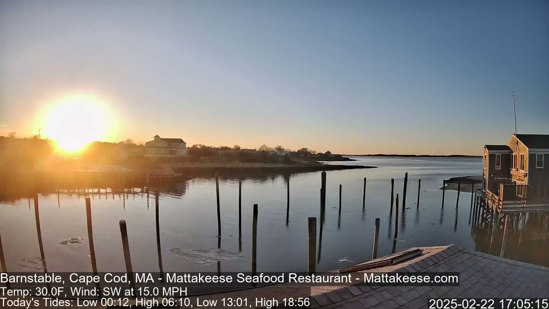 A calm body of water at sunset, reflecting the bright sun, with a few buildings visible on the shore and several posts in the water.