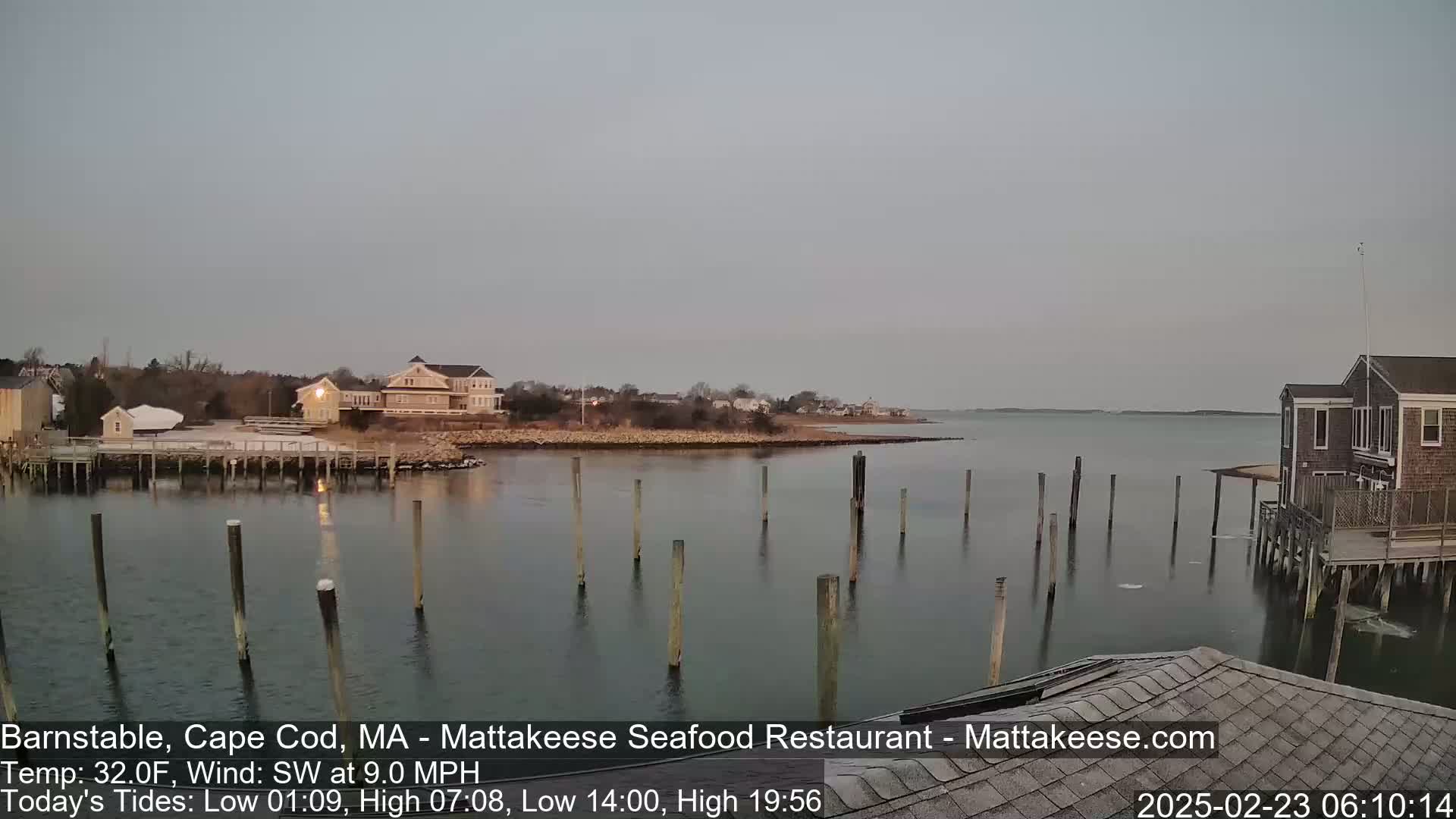 A calm body of water with wooden pilings, houses, and a shoreline under a light gray sky.