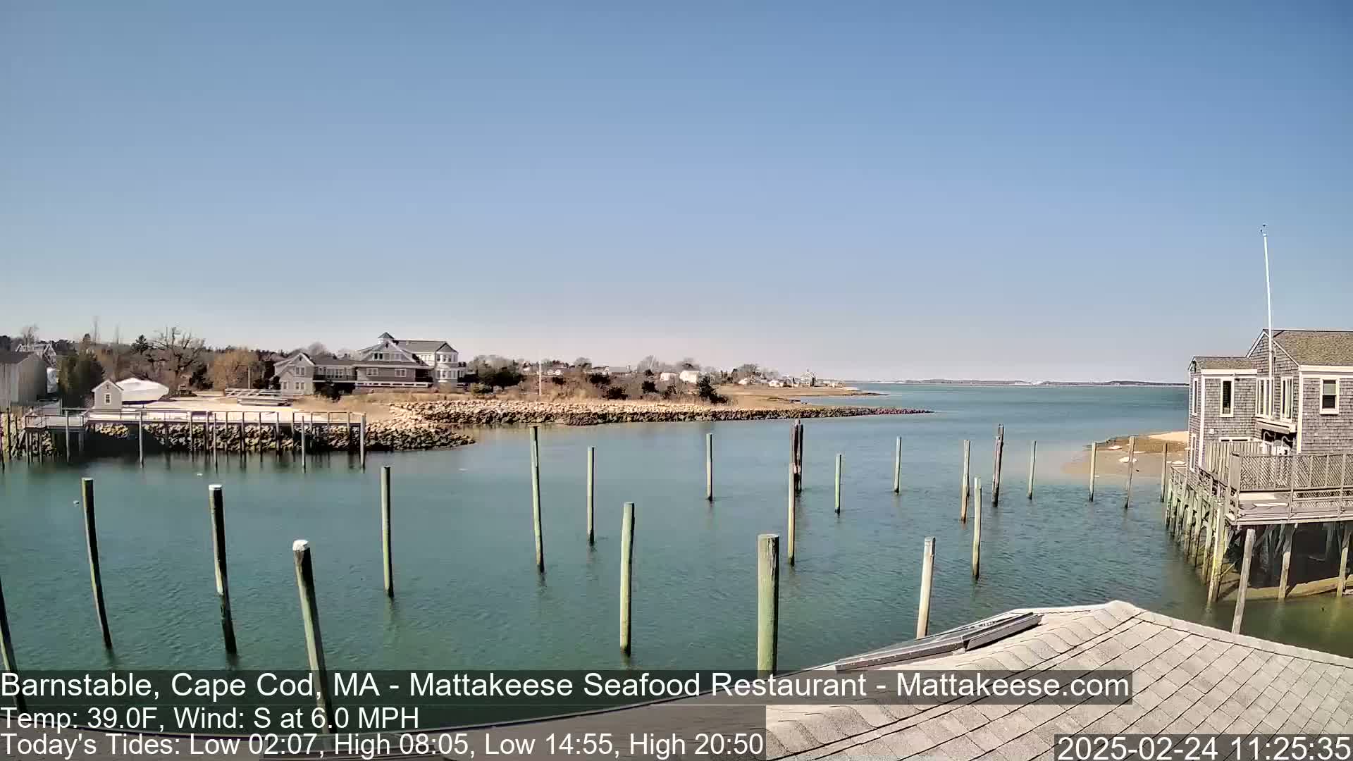 A calm, sunny day reveals a harbor scene with numerous pilings in the water, houses on the shore, and a clear view of the ocean beyond.