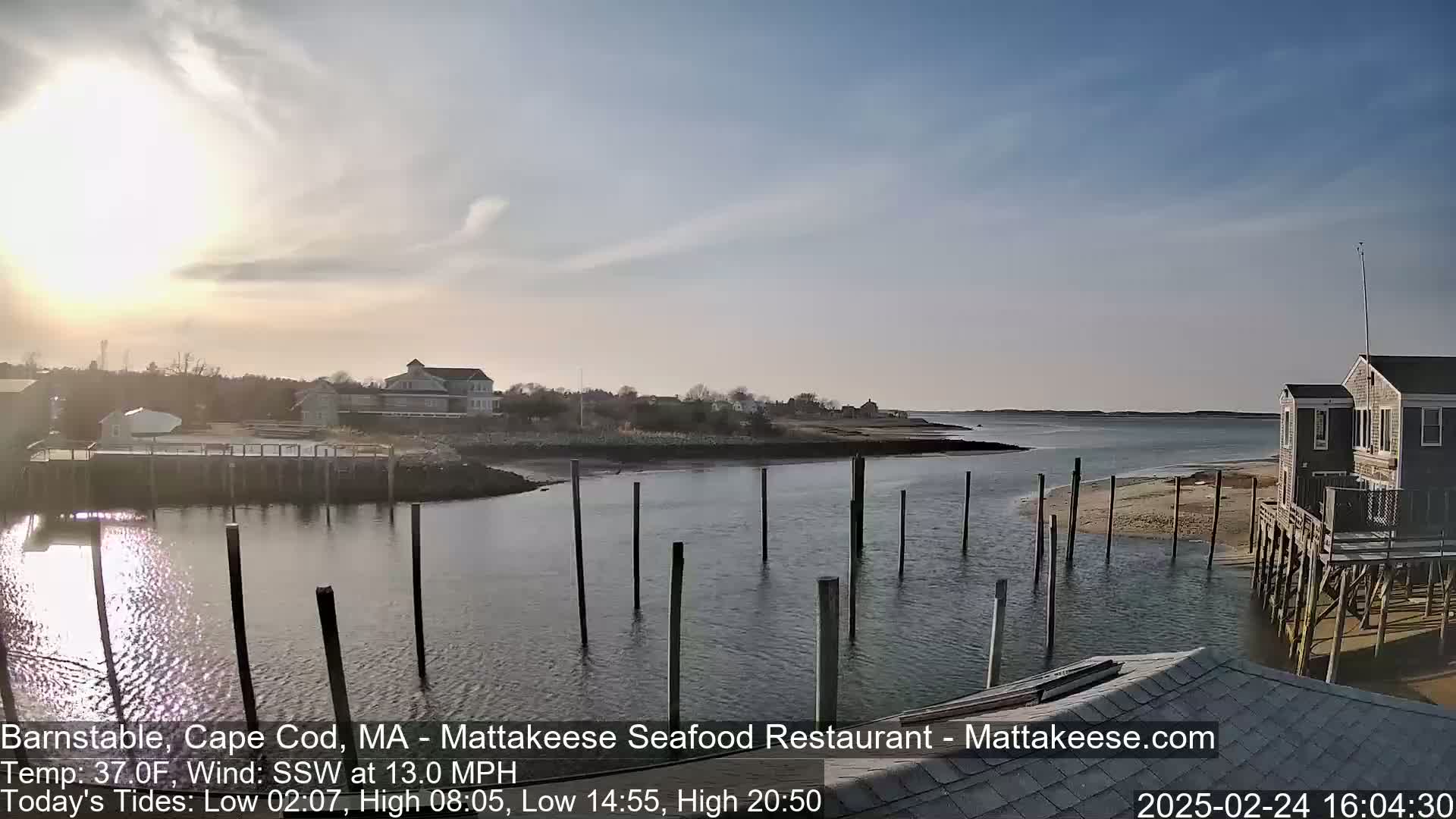 A calm, sunny day reveals a harbor scene with several buildings, a small sandy beach, and a row of pilings in the water.