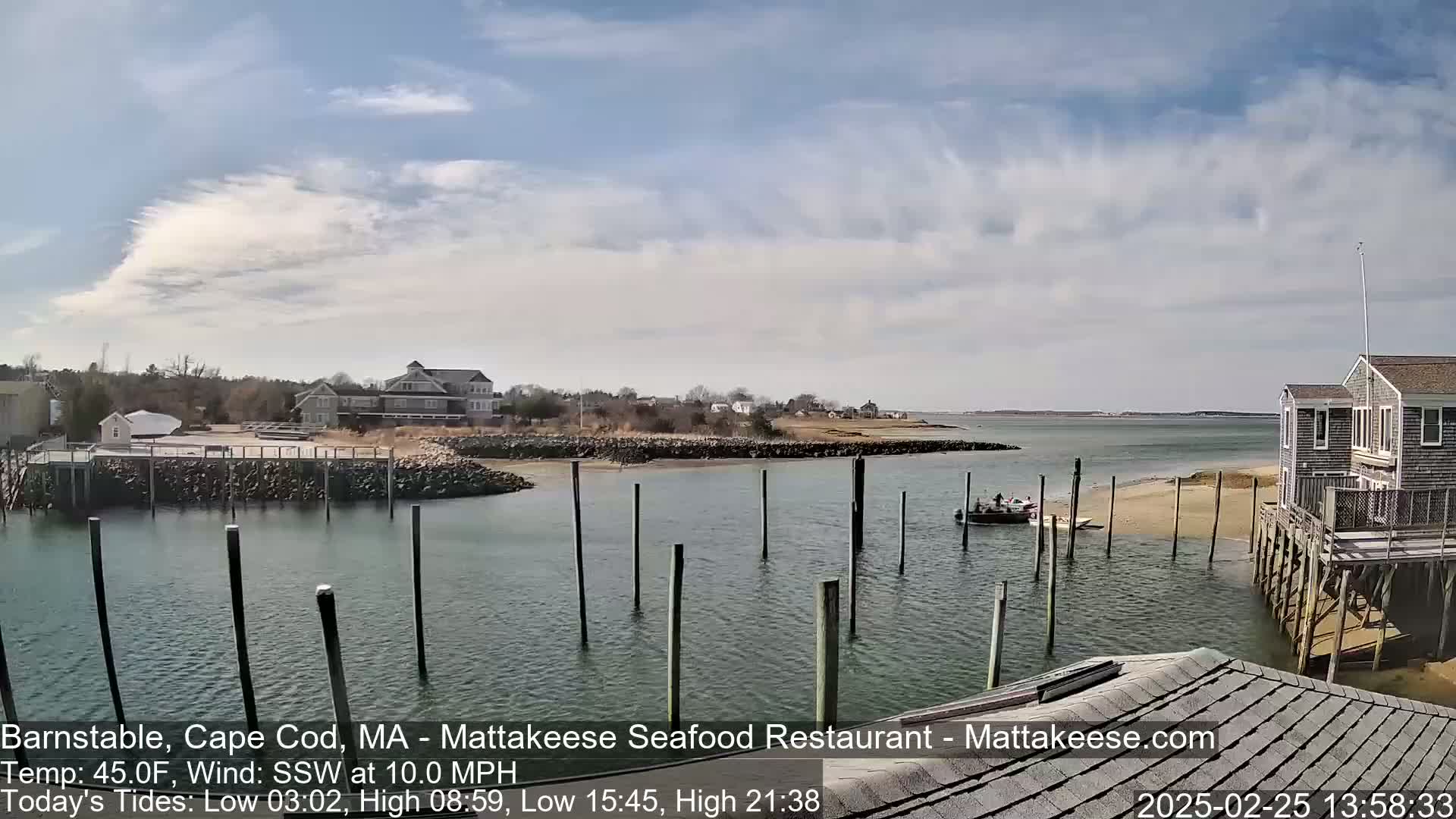 A partly cloudy day shows a calm harbor with boats and pilings, houses, and a sandy shore.