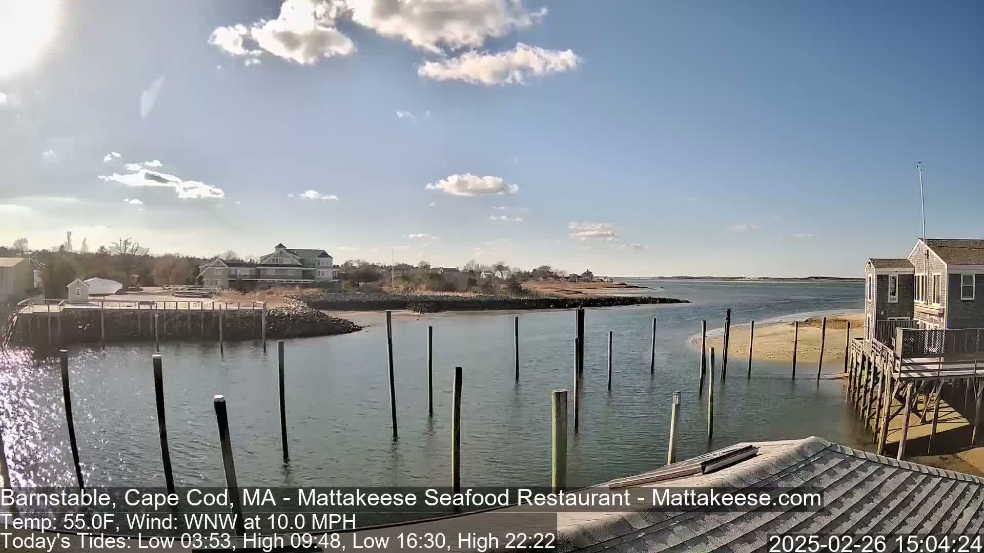 A calm, sunny day shows a waterfront scene with a calm body of water, wooden pilings in the water, buildings along the shore, and a partly cloudy sky.