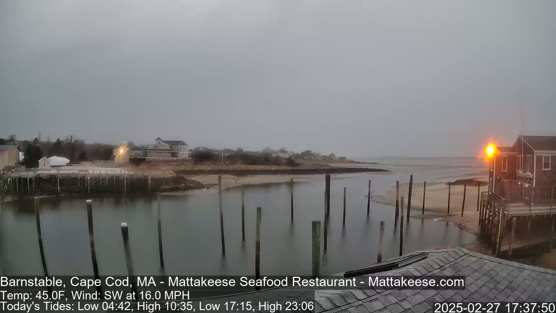 A calm, overcast day shows a view of a harbor with wooden pilings in the water, houses and buildings along the shore, and a sandy area at low tide.