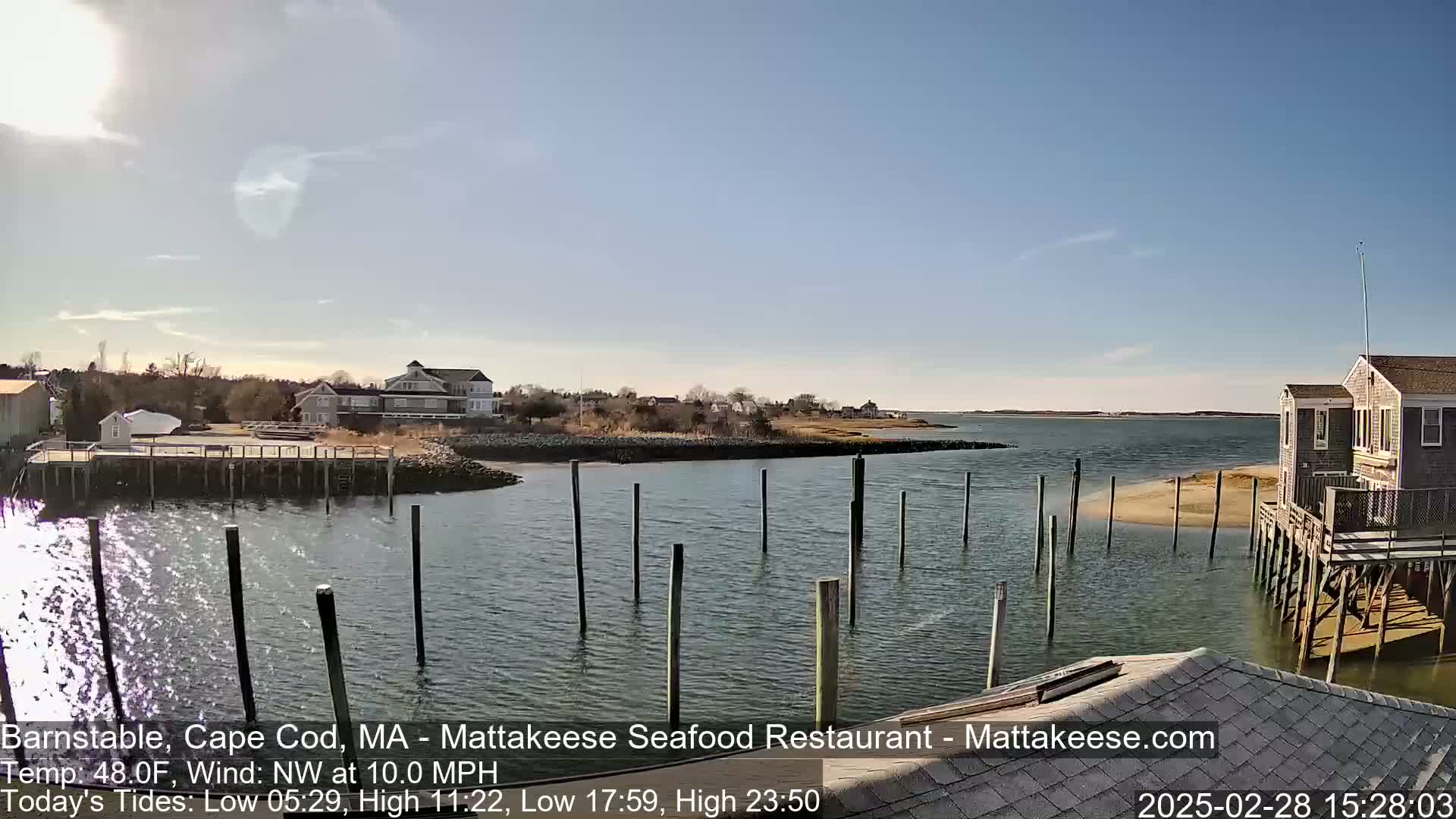 A waterfront scene on a sunny day shows a calm body of water with wooden pilings, a sandy shore, and houses along the shoreline.