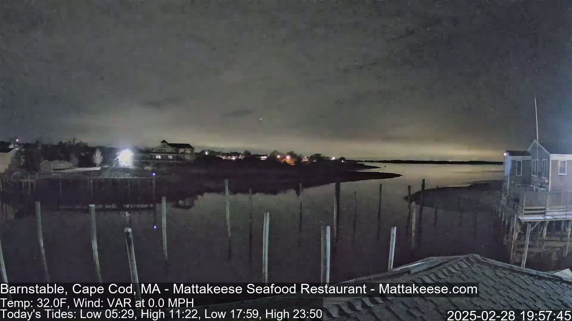 A nighttime view of a calm body of water, with a few buildings and pilings visible along the shoreline under a cloudy sky.