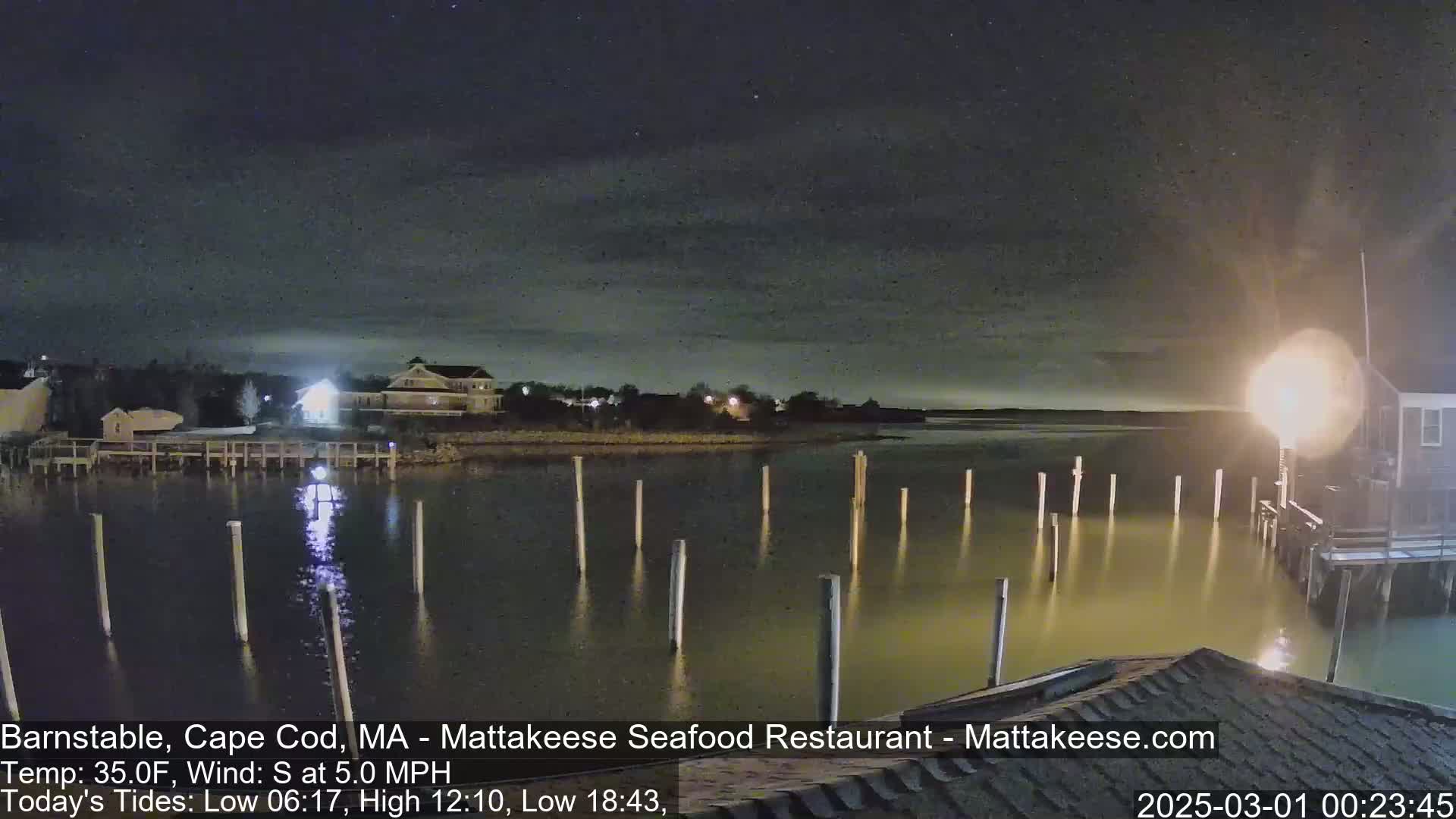 A nighttime view of a calm body of water with wooden pilings, houses on the shore, and a mostly clear sky.