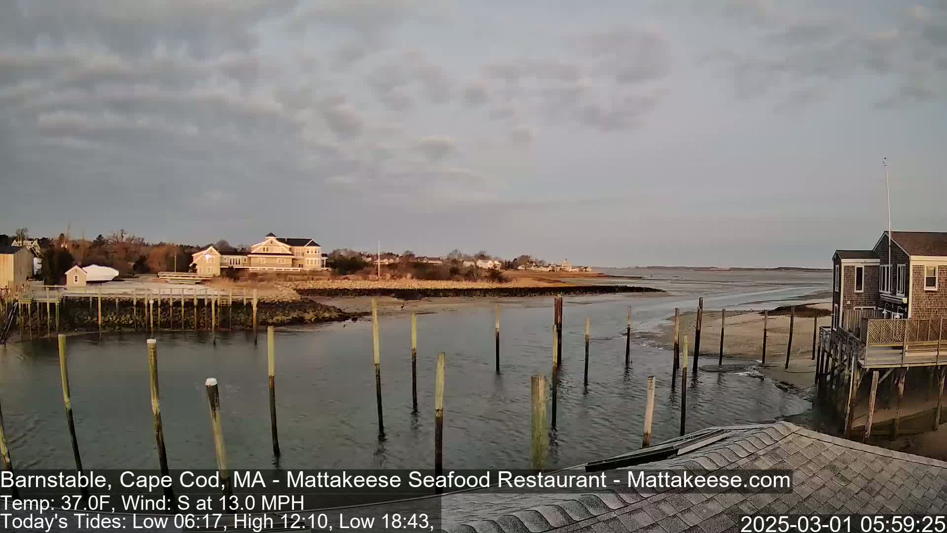A calm body of water with wooden pilings extends from a shoreline with houses, under a mostly cloudy sky.
