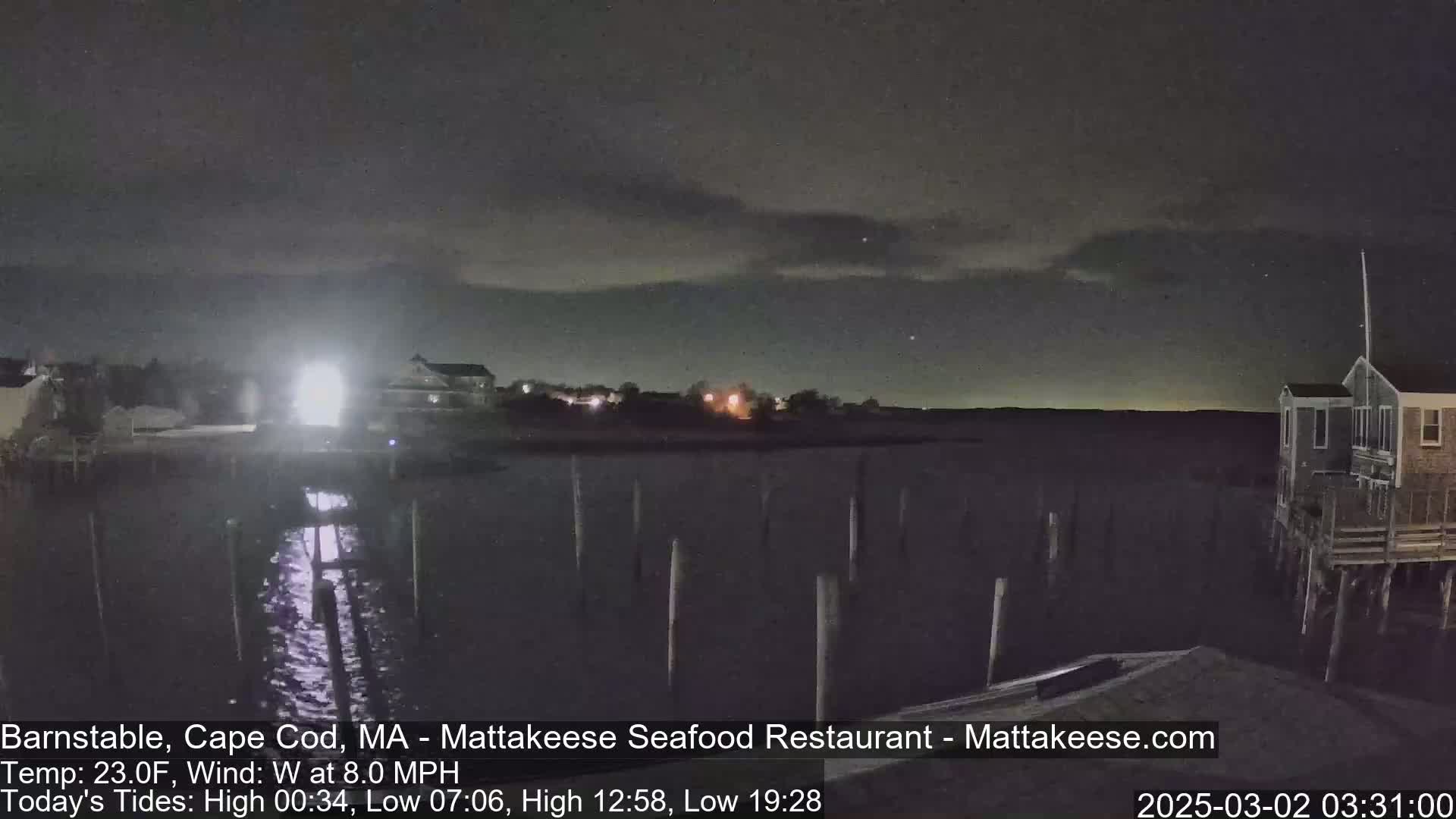 A nighttime view of a calm body of water with pilings and a few buildings visible under a partly cloudy sky.