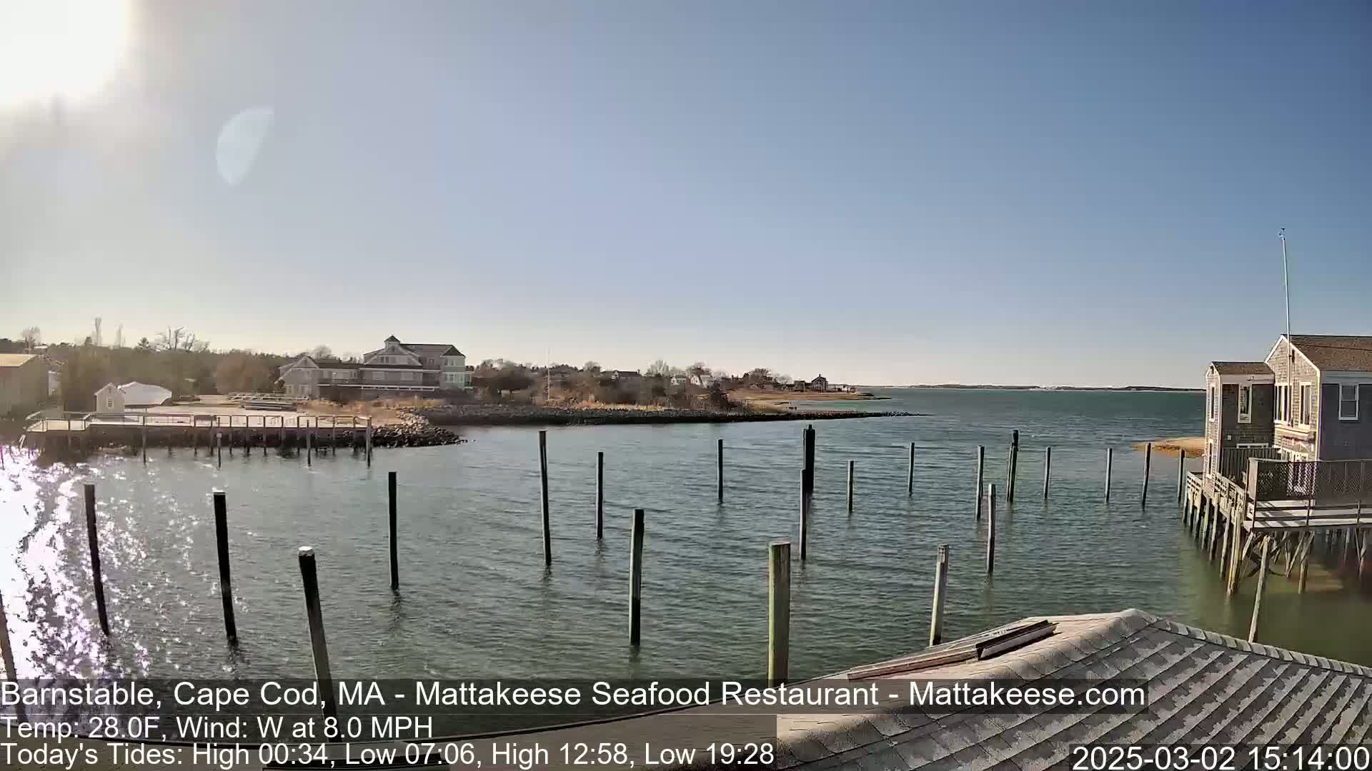 A sunny day shows a calm body of water with pilings extending into it, houses and buildings are visible on the shore, and a wooden structure is in the foreground.