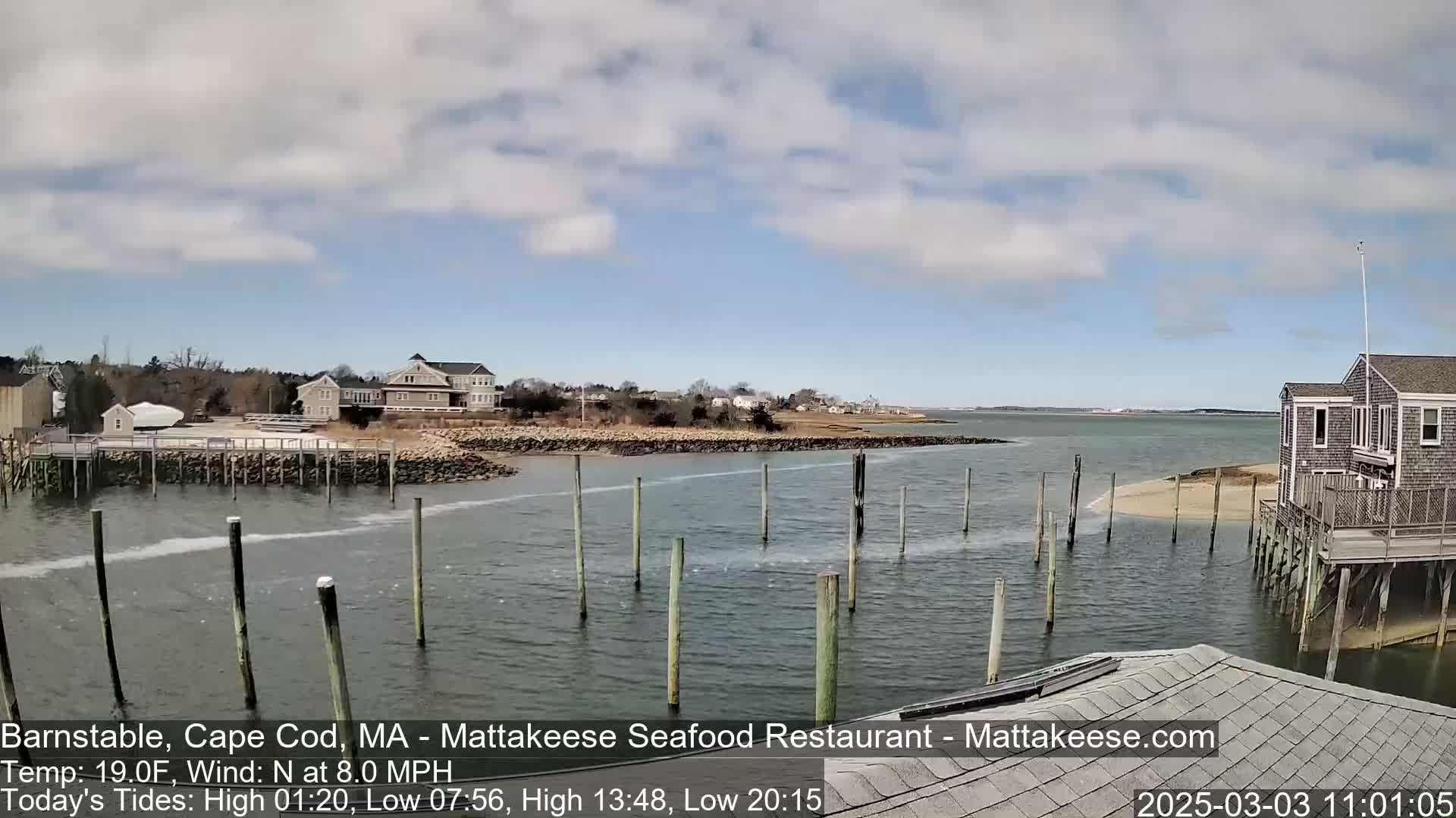 A partly cloudy day shows a calm body of water with wooden pilings in the foreground, a small sandy beach, and buildings along the shoreline.
