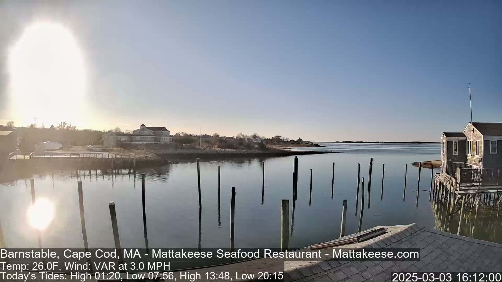 A sunny day reveals a calm body of water with wooden pilings extending into it, buildings on the shore, and a bright sun partially obscured by atmospheric haze.