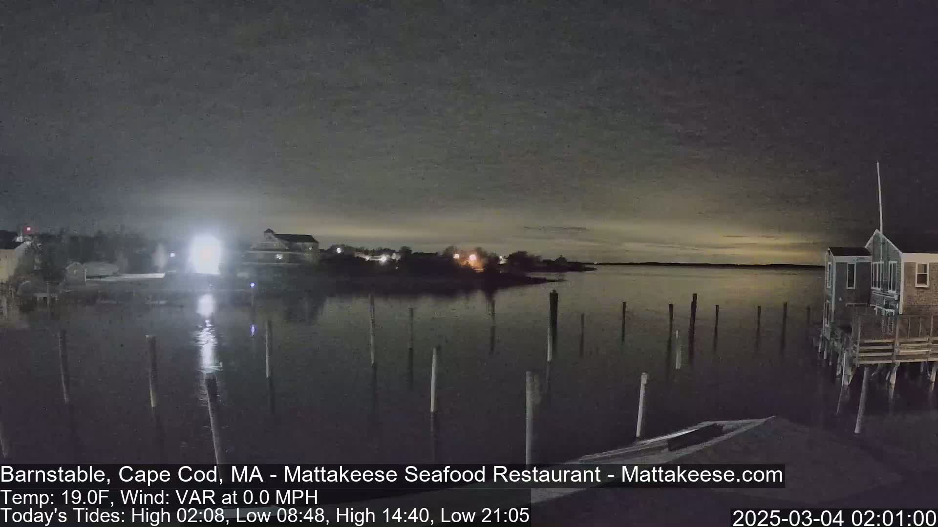A nighttime view of a calm body of water with several pilings and buildings along the shore under a dark, mostly clear sky.