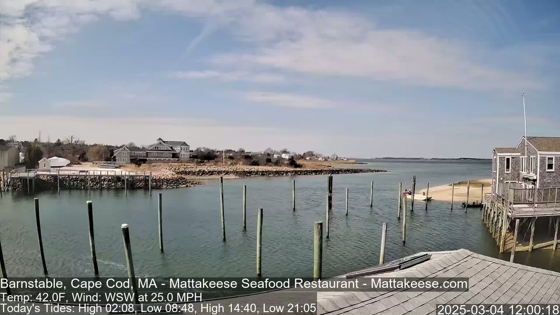 A calm, partly cloudy day shows a coastal scene with a small harbor, houses, and a restaurant on stilts over the water.