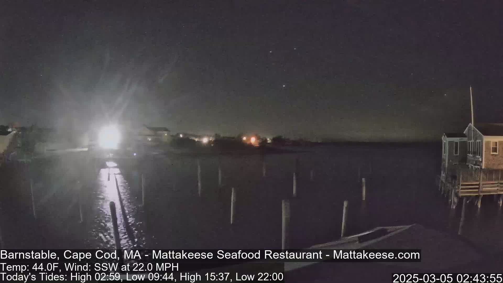 A nighttime view of a waterfront scene with a stormy, windy atmosphere shows a dark body of water with pilings, a building on stilts, and distant lights under a partly cloudy sky.