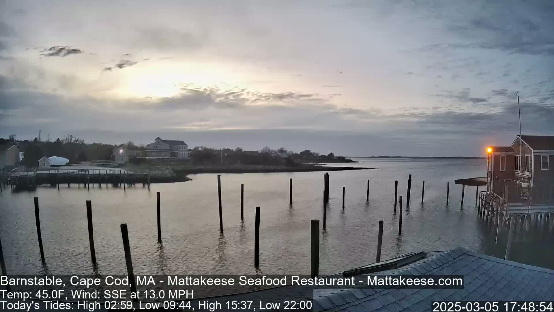 A calm body of water with numerous pilings extends from a shoreline featuring buildings and low vegetation under a partly cloudy, dusky sky.