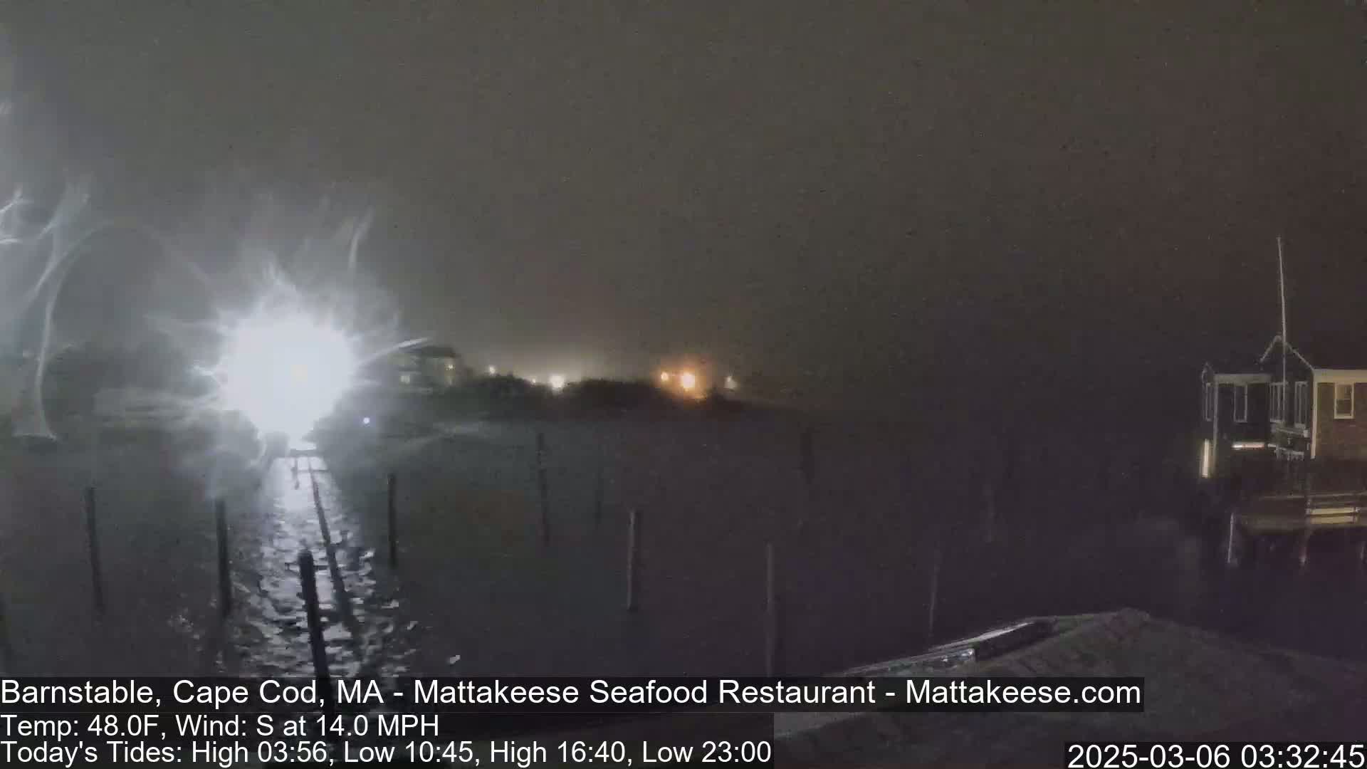 A nighttime scene of a waterfront area with heavy snow or blowing snow, illuminated by distant lights.