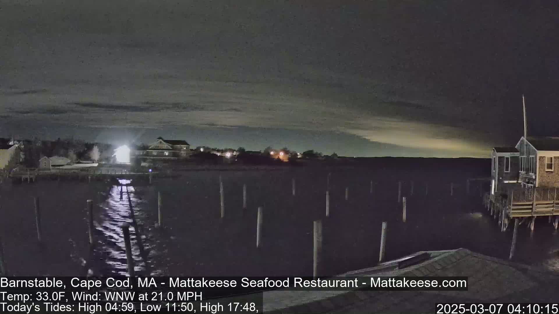 A nighttime view of a calm body of water with pilings, buildings along the shore, and a faintly lit sky.