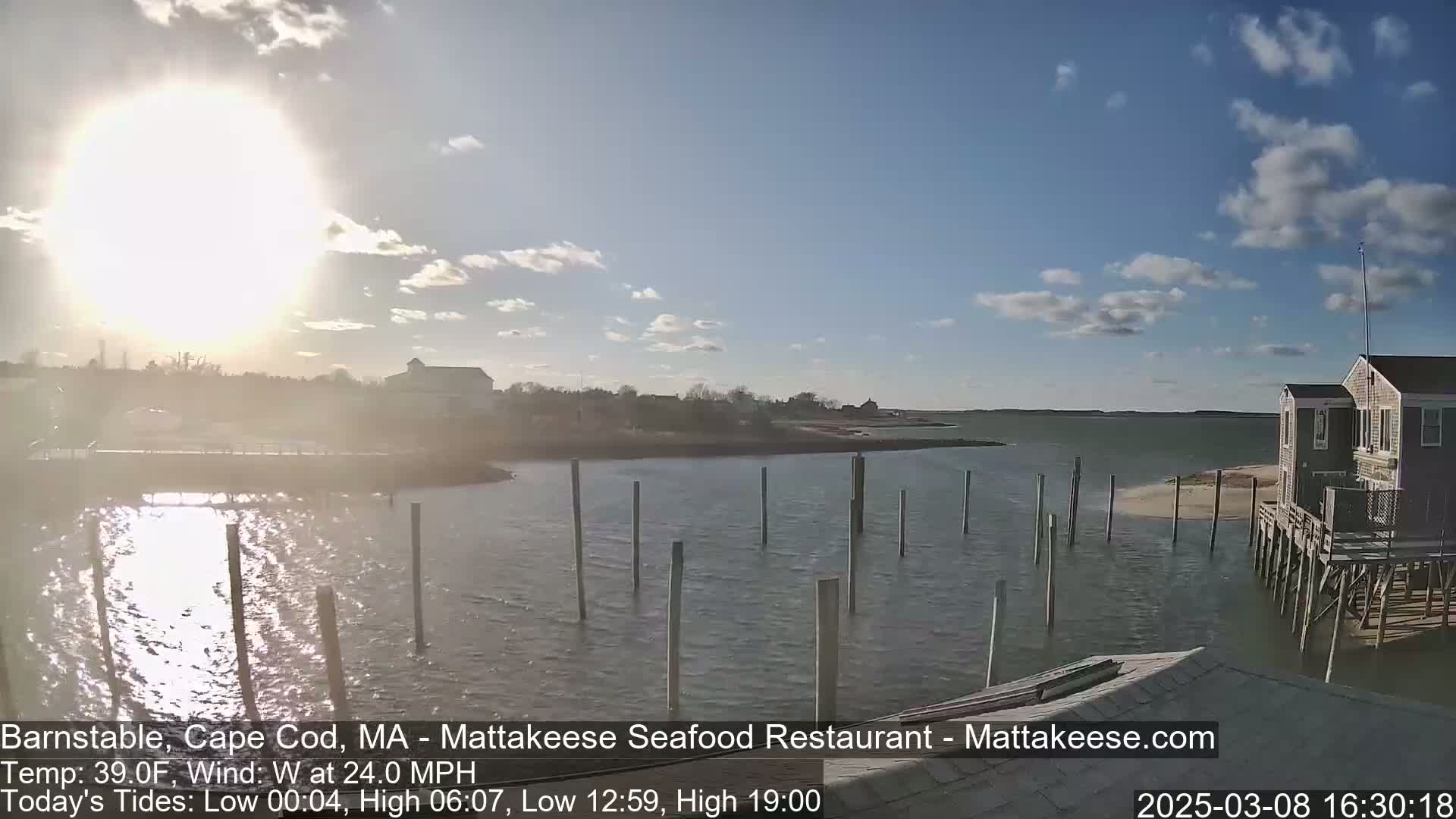 A bright, sunny day with a strong westerly wind shows a calm body of water with numerous pilings and a small house on stilts in the foreground, and a shoreline in the background.