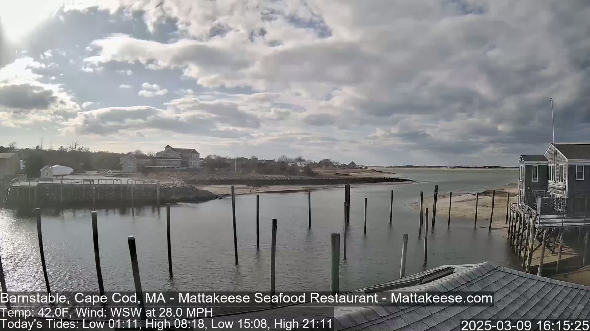 A partly cloudy day shows a calm body of water with wooden pilings and buildings along the shore.