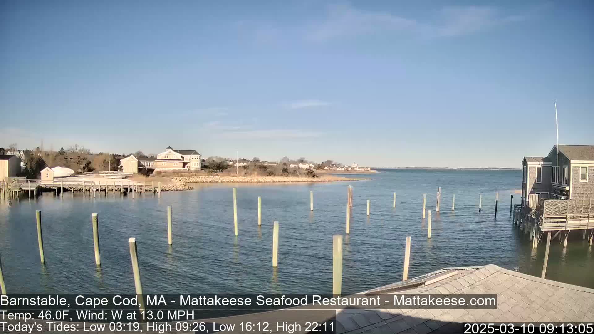 A calm, sunny day shows a waterfront scene with buildings, a small dock with posts in the water, and a body of water extending to a distant shoreline.