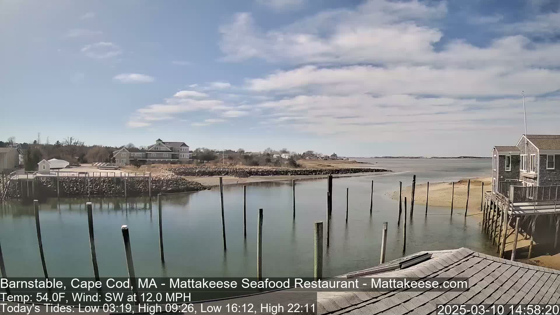 A calm body of water with wooden pilings, a sandy shore, and several buildings in the background under a partly cloudy sky.