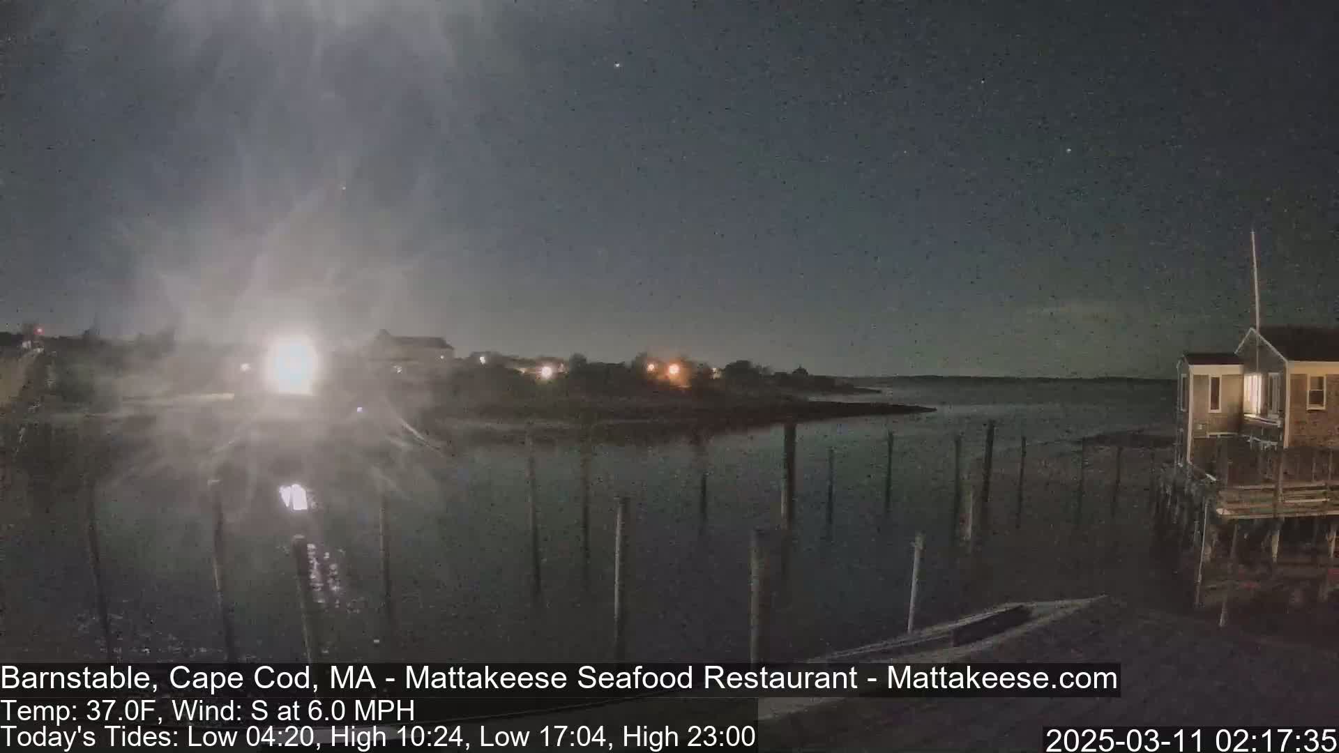 A nighttime view of a calm body of water with pilings extending into it, a building on stilts to the right, and dimly lit buildings and land in the distance under a dark, clear sky.