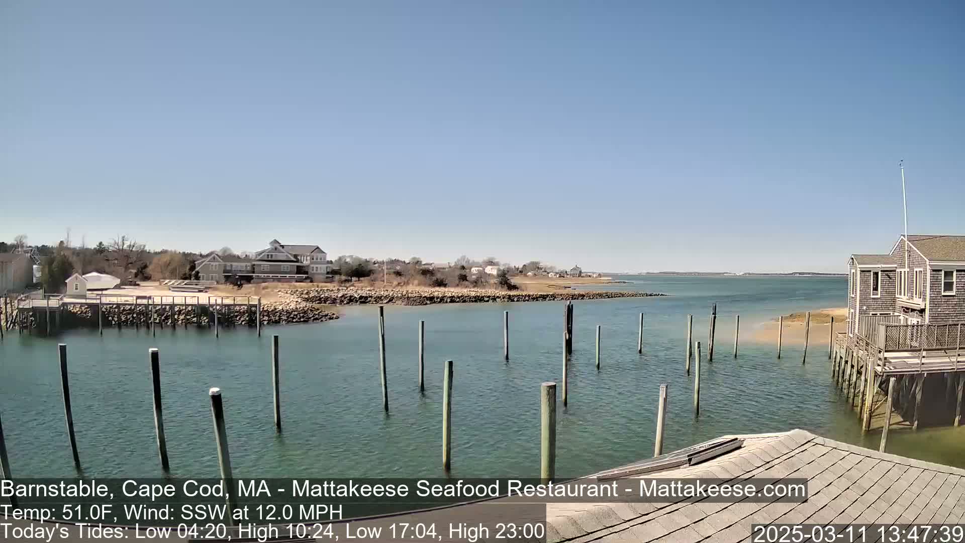 A calm, sunny day shows a waterfront scene with a calm body of water, wooden pilings, several buildings, and a sandy shoreline.
