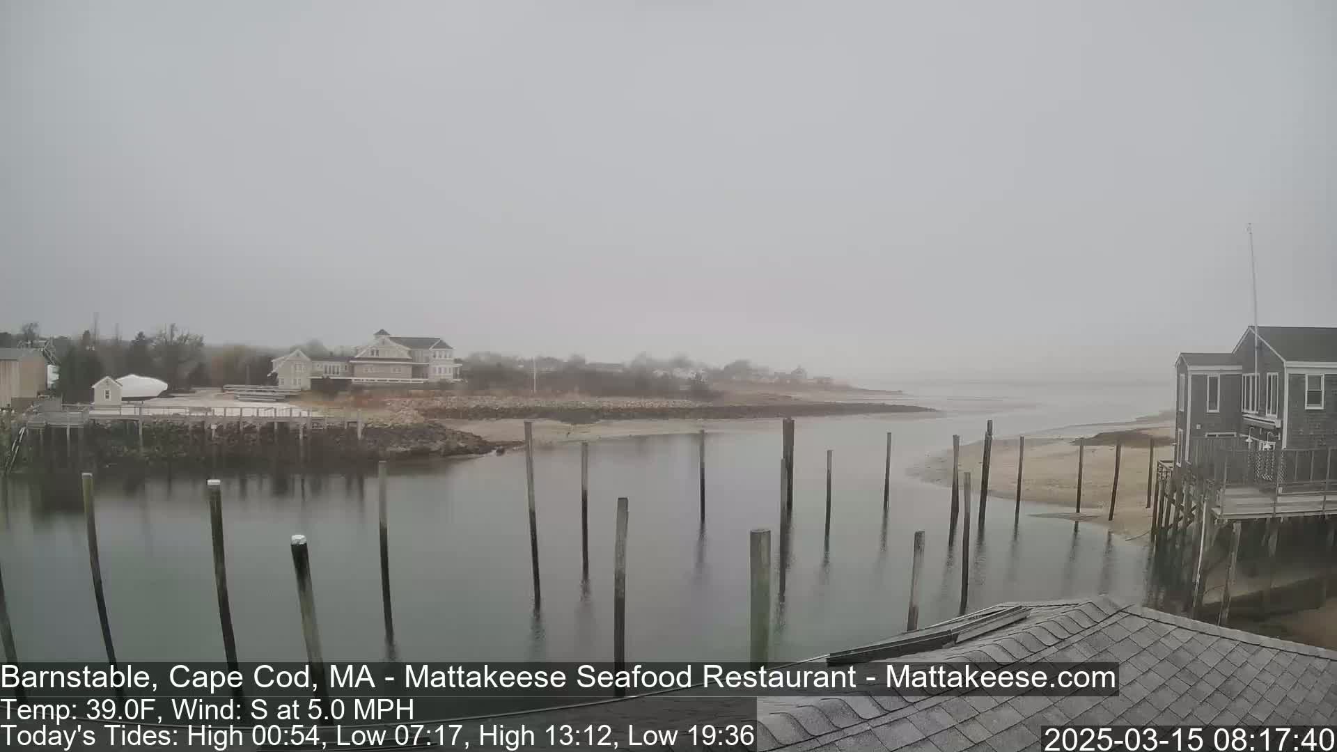 A foggy coastal scene shows a calm body of water with wooden pilings, houses, and a sandy shore.
