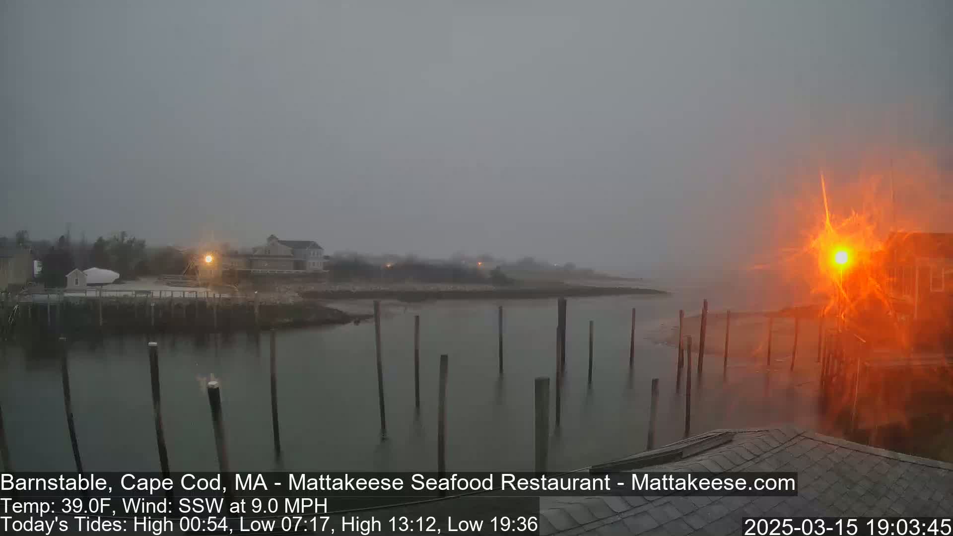 A foggy, windy scene of a harbor with wooden pilings, buildings, and a bright orange light in the foreground.