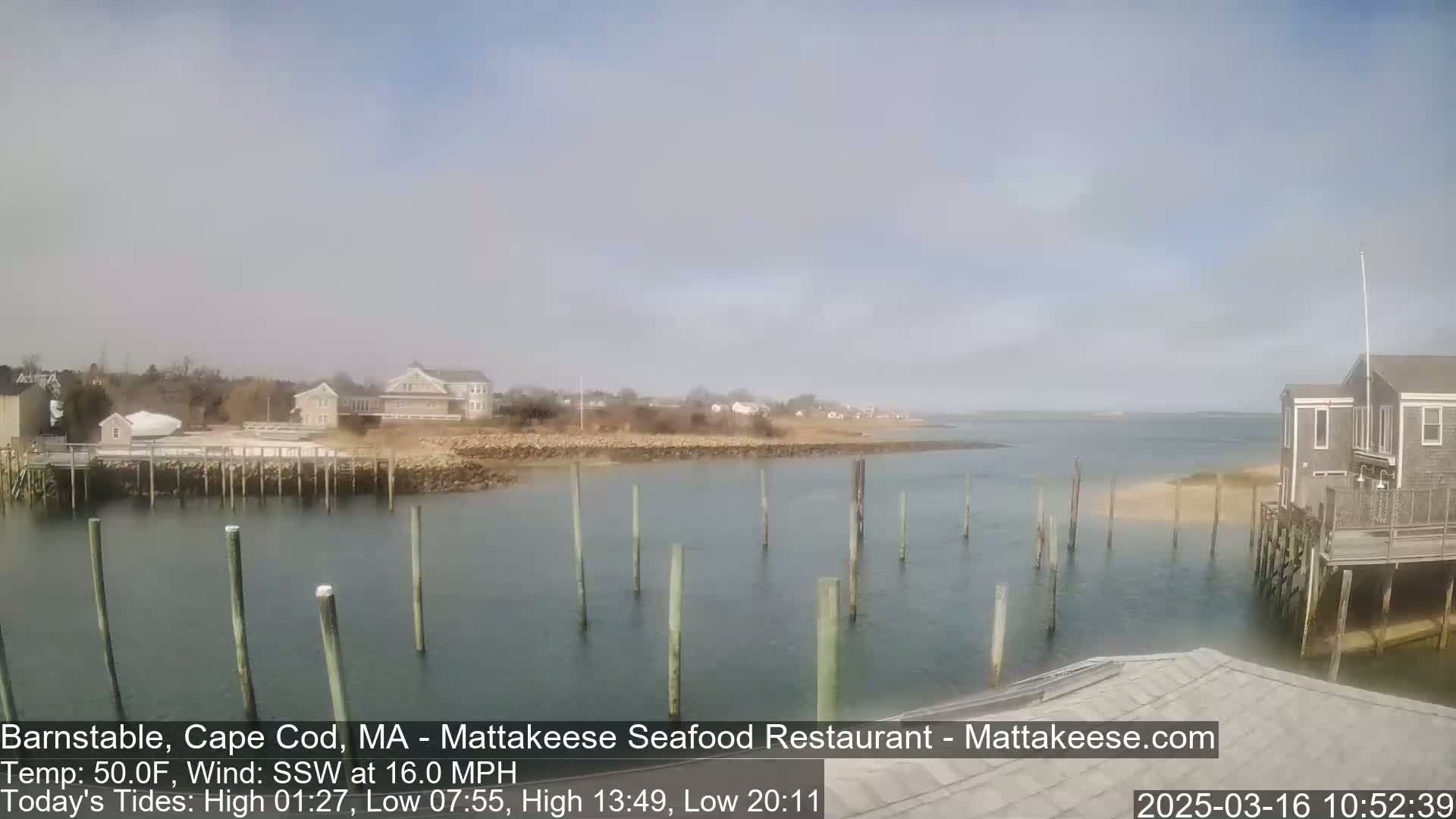 A calm, partly cloudy day shows a coastal scene with a harbor, wooden pilings, and buildings along the shore.