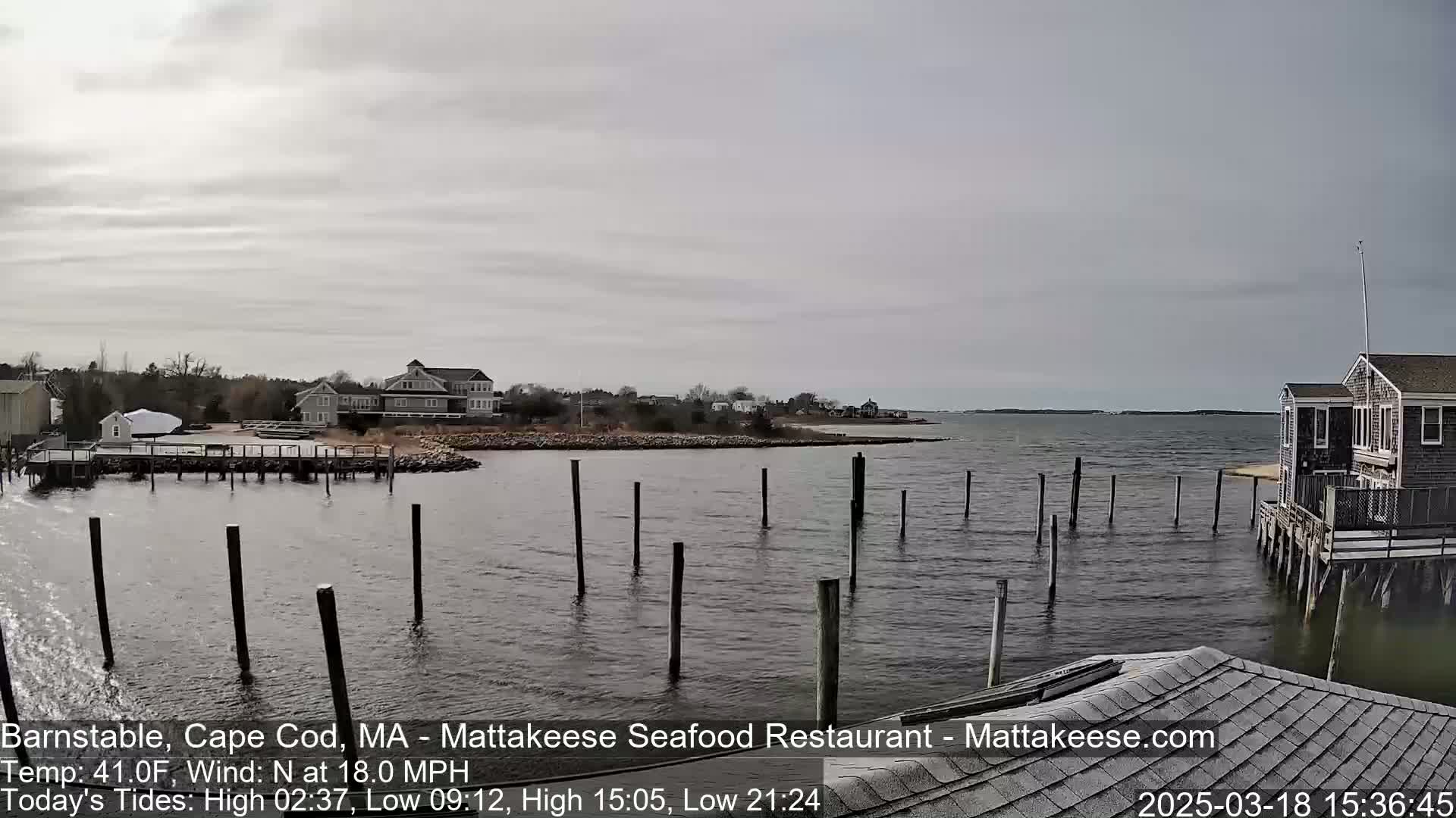 A calm, gray day shows a waterfront scene with several houses, a pier, and a small building on pilings extending into a body of water under an overcast sky.