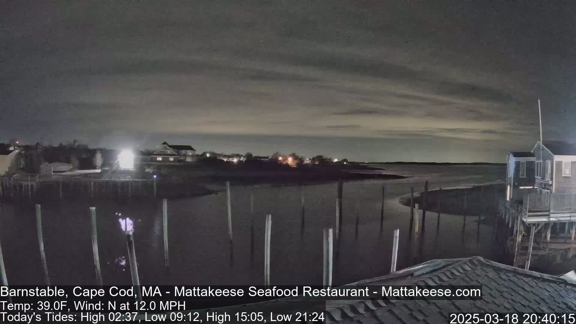 A nighttime view of a calm body of water, with a few buildings and pilings visible under a mostly cloudy sky.