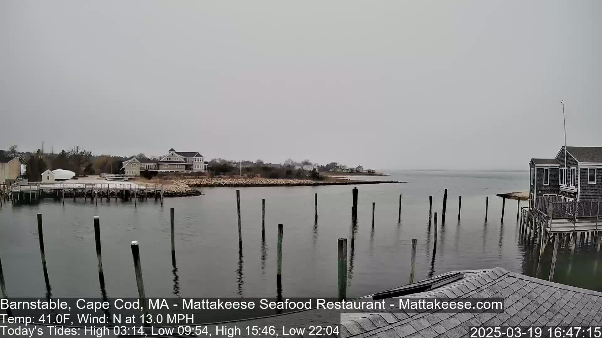 A calm, overcast day shows a waterfront scene with a calm body of water, wooden pilings, and houses along the shore.