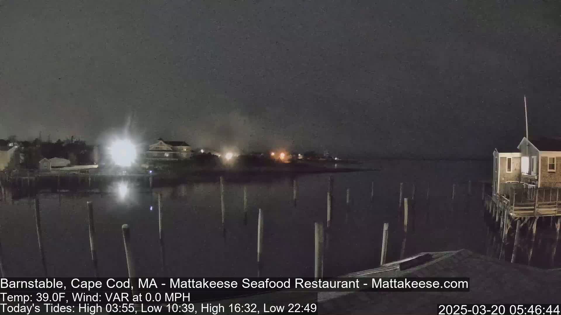 A nighttime view of a calm body of water with pilings, a few buildings on the shore, and some distant lights under a dark sky.