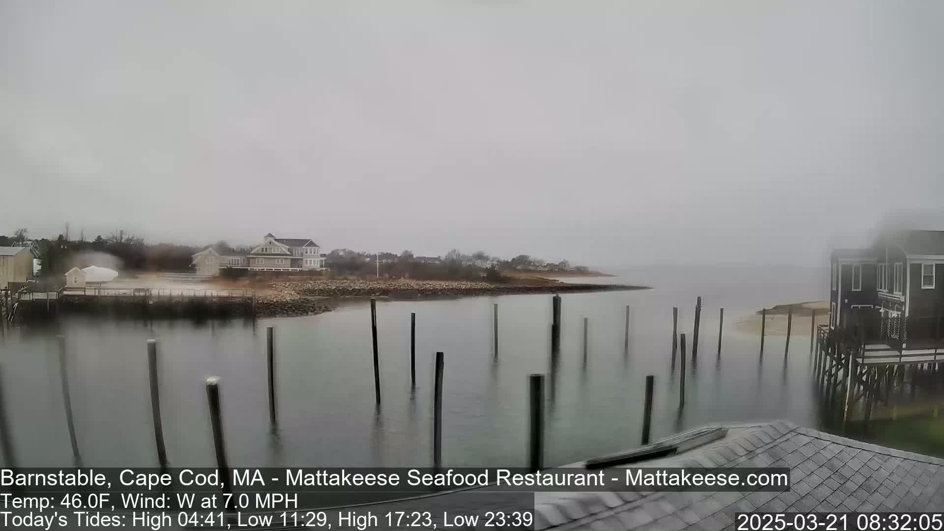 A foggy, overcast scene shows a calm body of water with several pilings extending from the shore, houses on the shore, and a bit of land extending into the water.