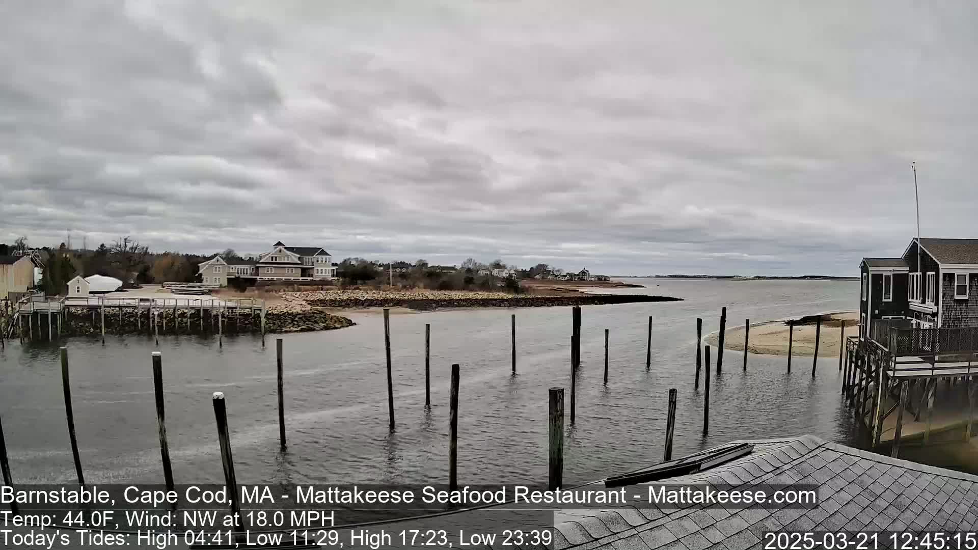 A calm bay with wooden pilings in the water, under a cloudy sky, is viewed from a waterfront home.