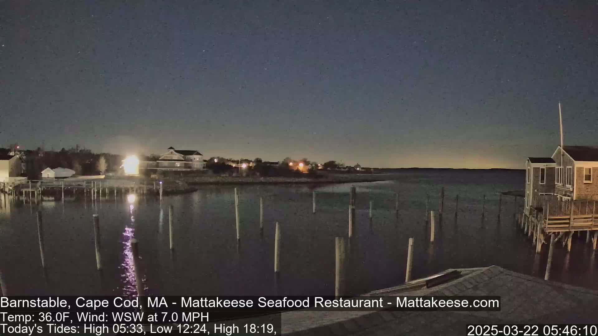 A nighttime view of a calm body of water with a few buildings and pilings along the shore under a clear, dark sky.
